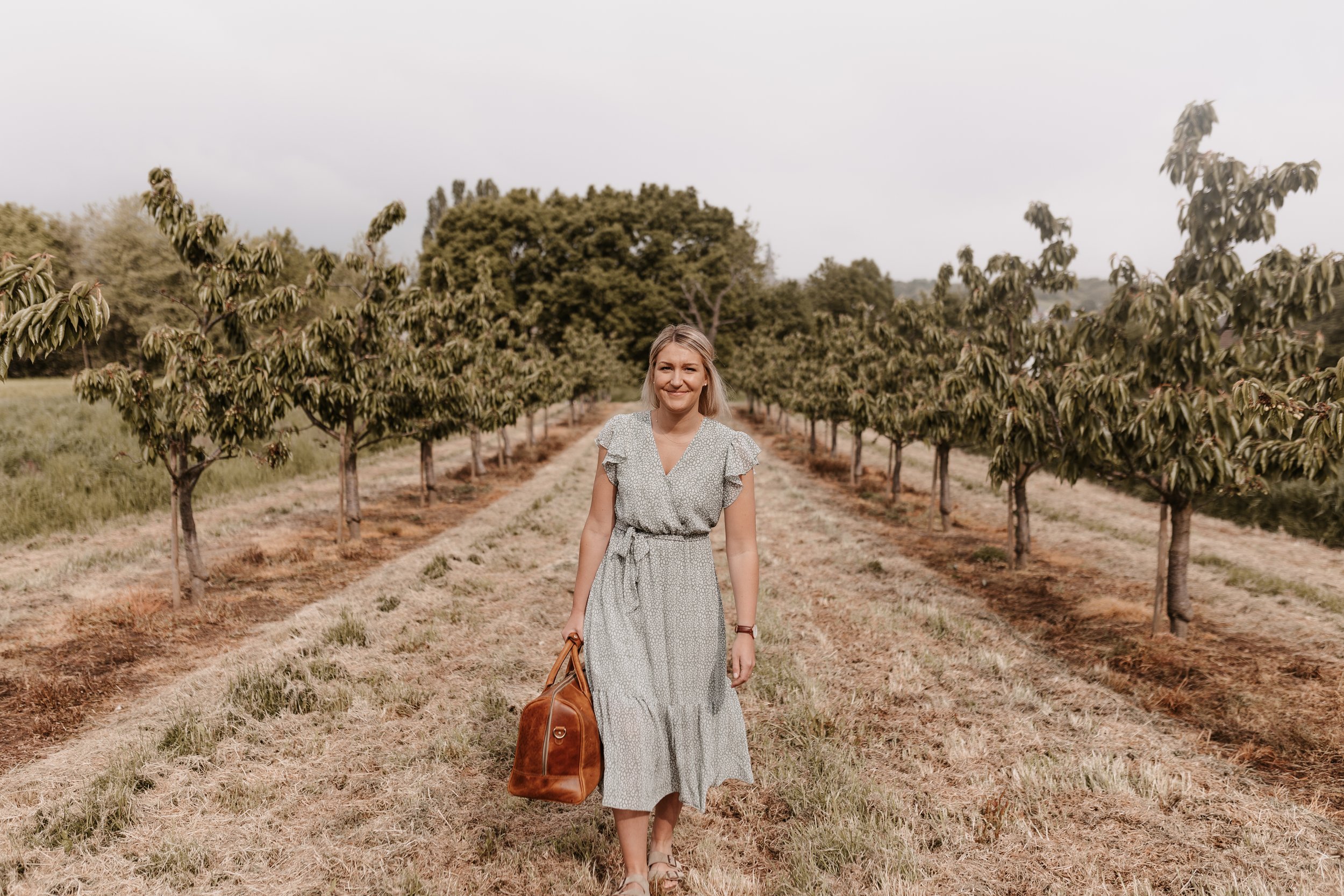 Eine junge Frau in einem Sommerkleid läuft zwischen Reihen von Obstbäumen, trag eine braune Tasche in der Hand, im Freien bei bewölktem Himmel.