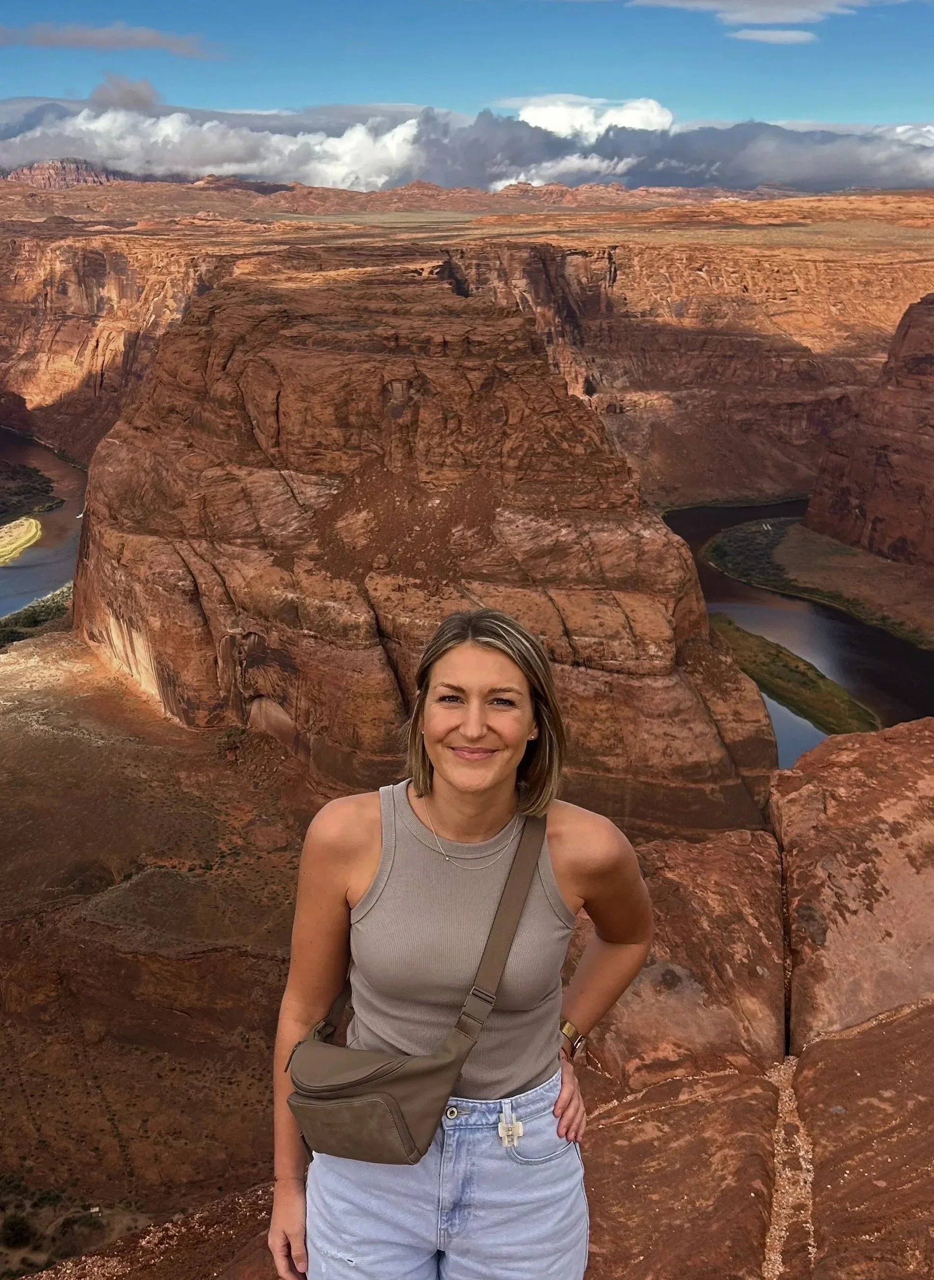 Eine junge Frau steht auf einem roten Felsen in einer Wüstenlandschaft mit Canyon und Fluss im Hintergrund. Die Landschaft ist trocken, mit hohen, roten Felsen und Gebirgen unter einem bewölkten Himmel.