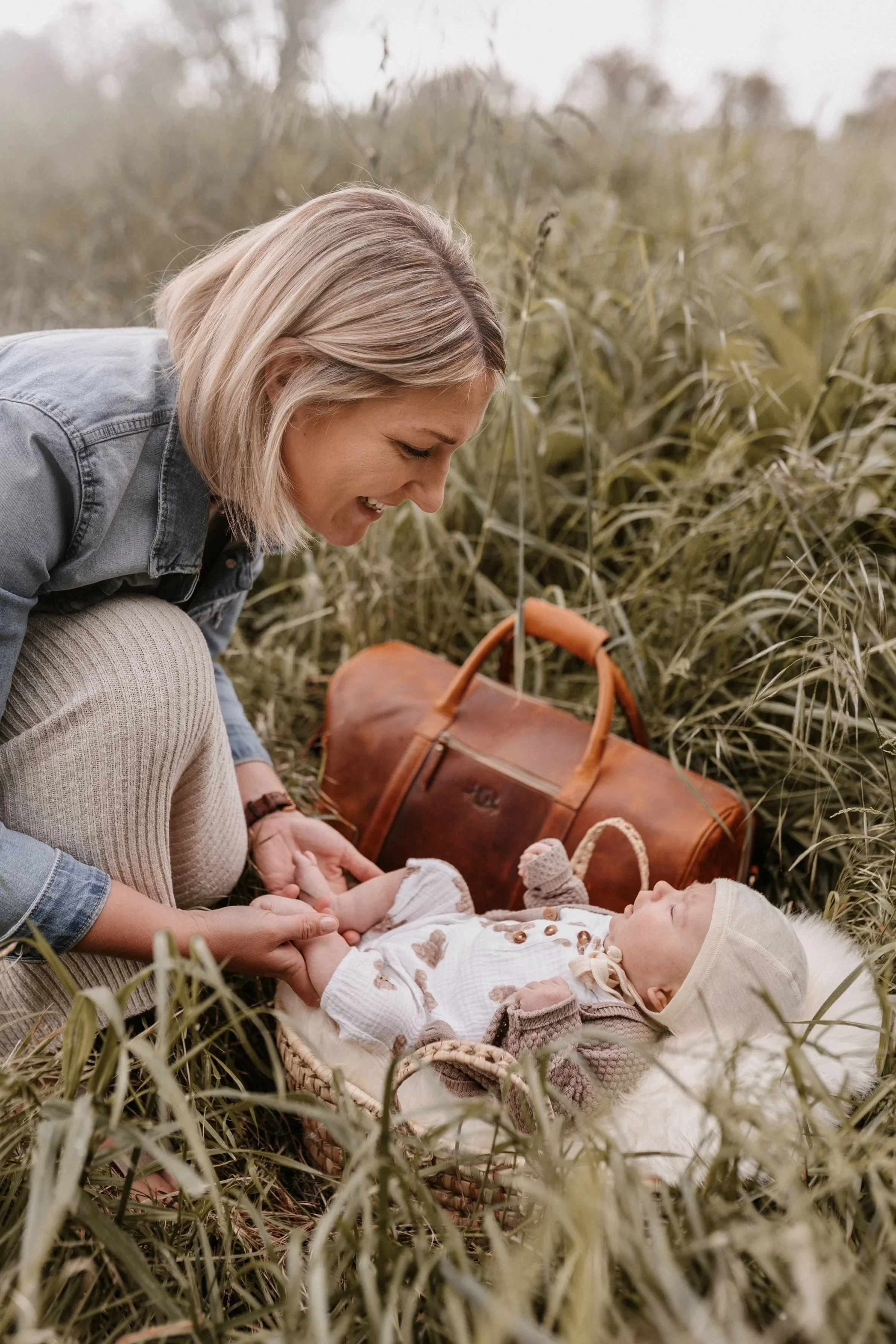 Eine Frau liegt in einem Feld mit Gras, hält die Hände eines schlafenden Babys, das in einem Korb liegt und trägt ein Cremefarbenes Outfit mit Hut, neben einem braunen Lederkoffer.