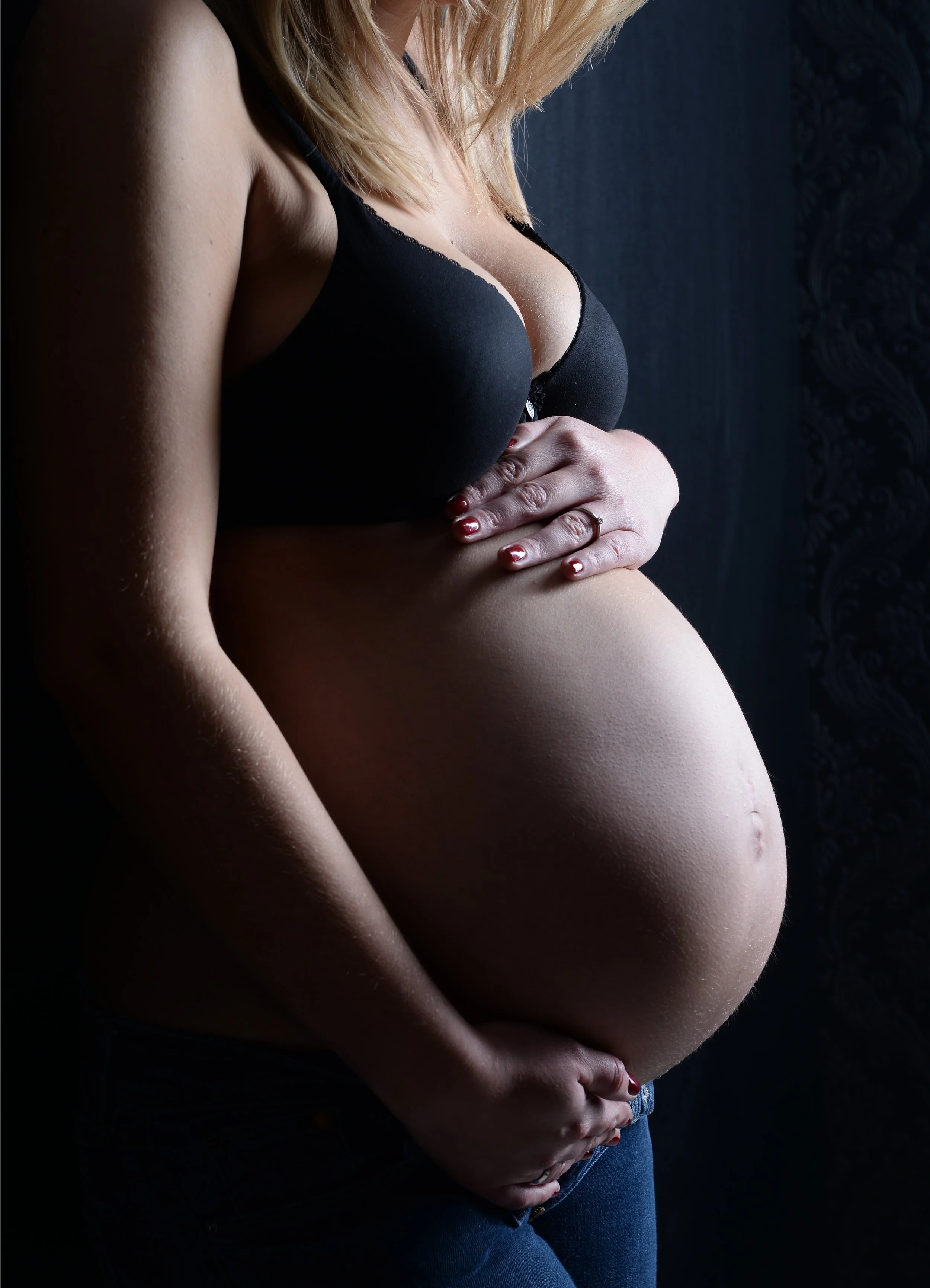 Schwangere Frau trägt schwarze Unterwäsche und hält ihren Babybauch, mit dunkelblauem Hintergrund und weichen Lichtverhältnissen.