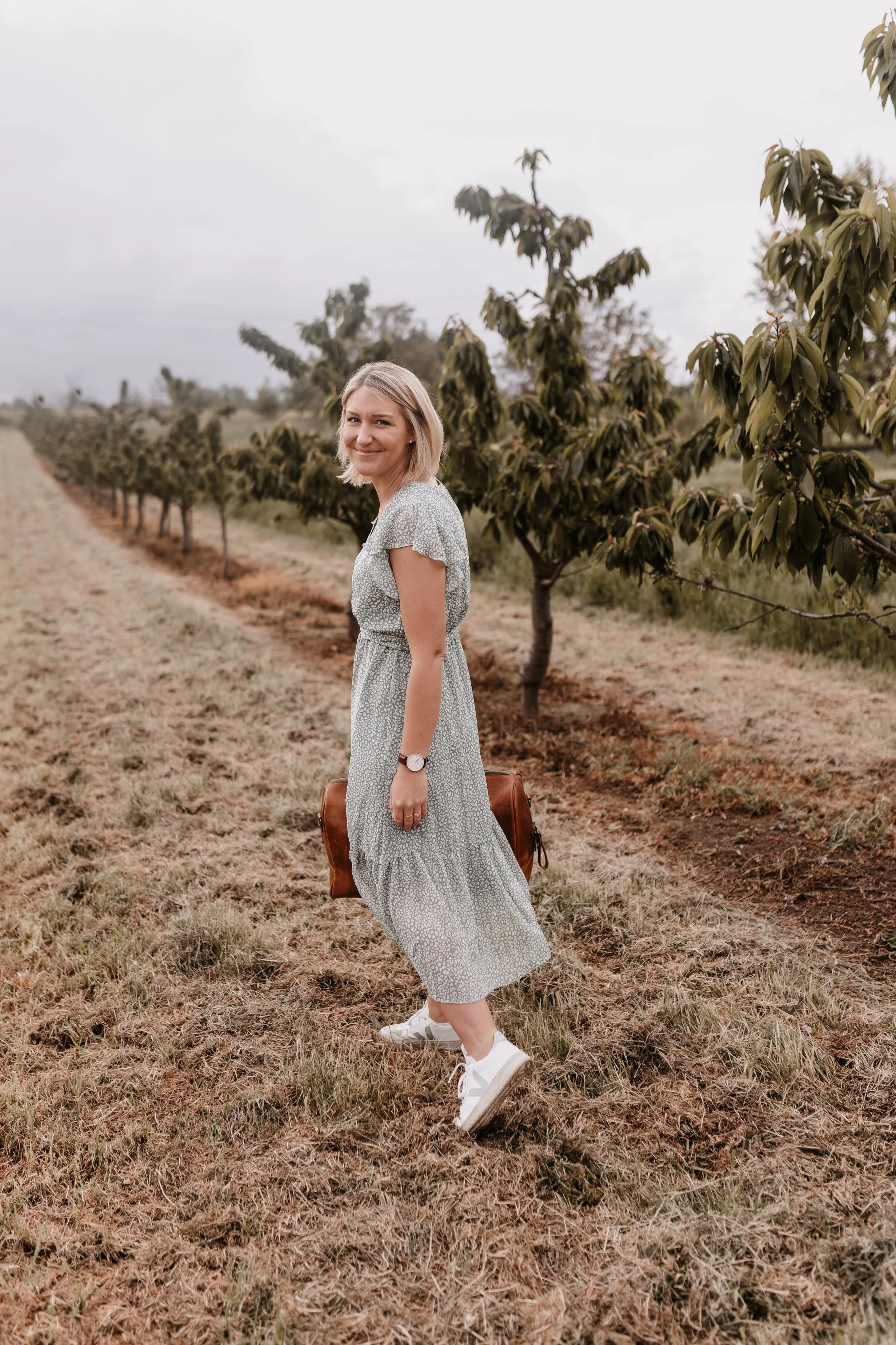 Eine junge Frau in einem Sommerkleid, weißen Turnschuhen und mit einer Armbanduhr steht in einem Obstgarten bei bewölktem Himmel und lächelt in die Kamera.