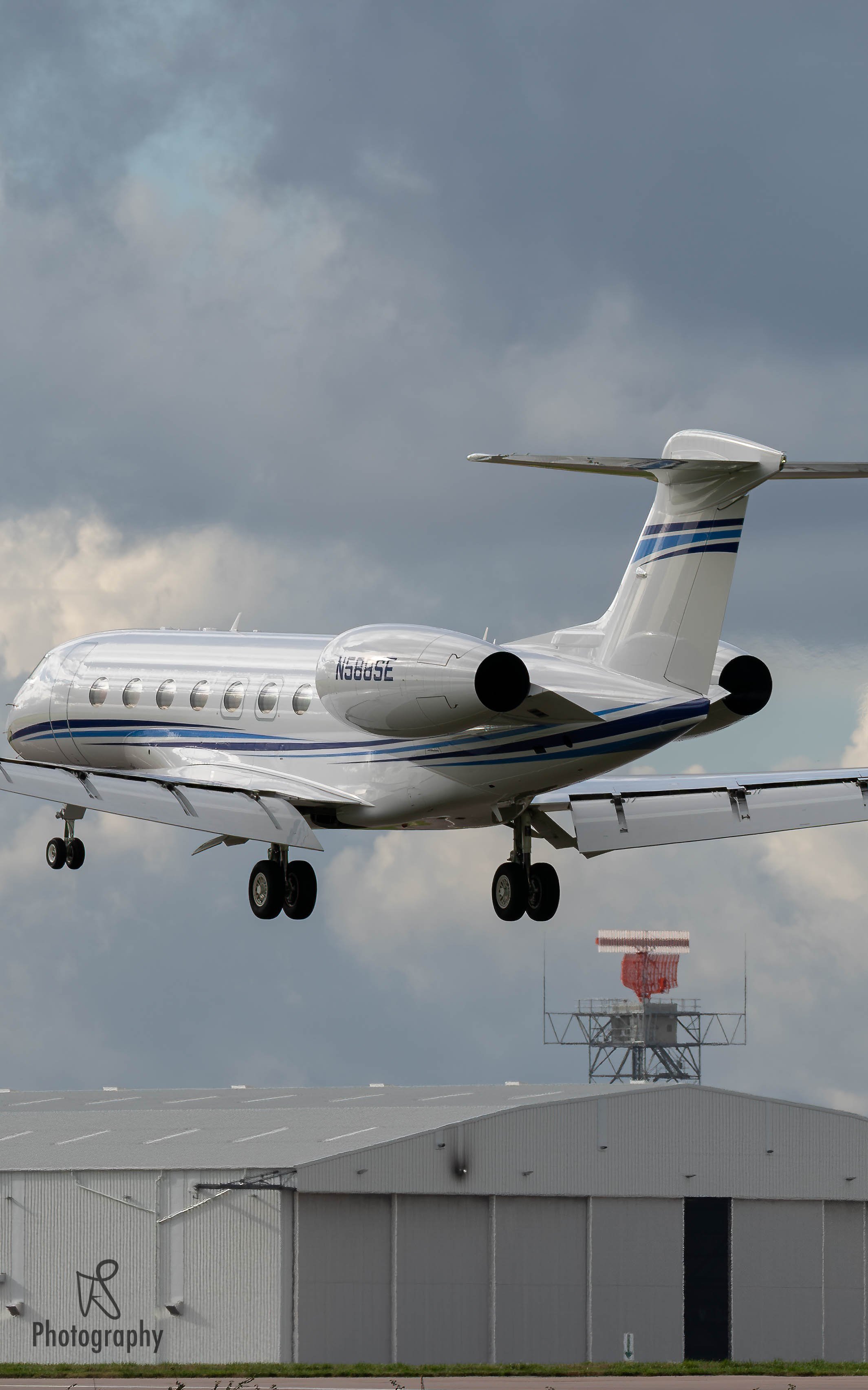 Private jet flying above a hangar with cloudy sky in the background.