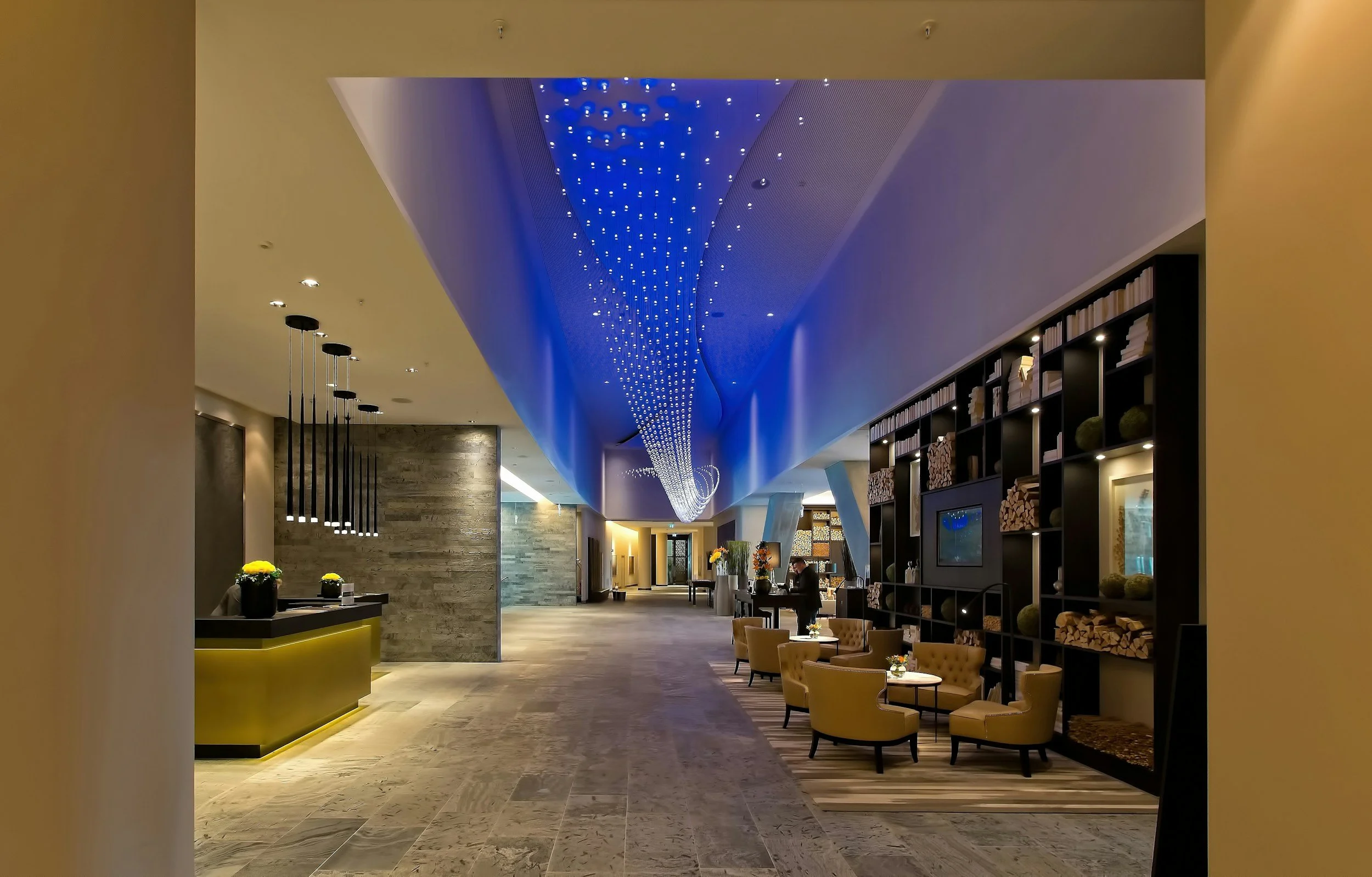 Modern hotel lobby with a high ceiling, blue LED lighting, seating area with yellow chairs, a reception desk, and decorative shelves with wood logs and ornaments.