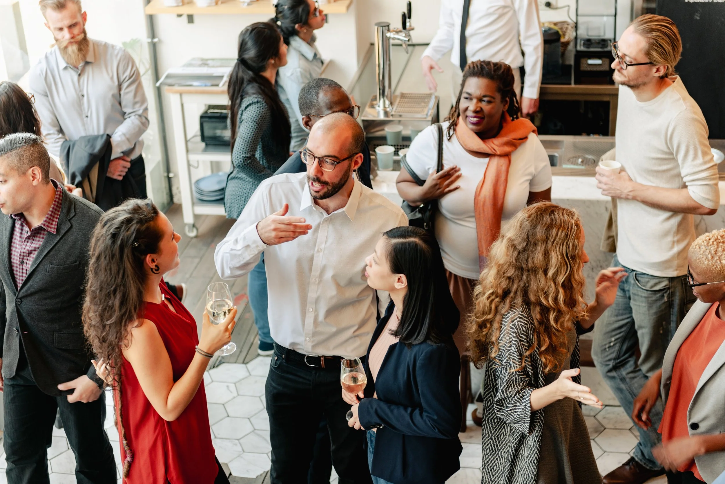 People socializing at a gathering in a kitchen or cafe setting, with some holding drinks and engaging in conversation.