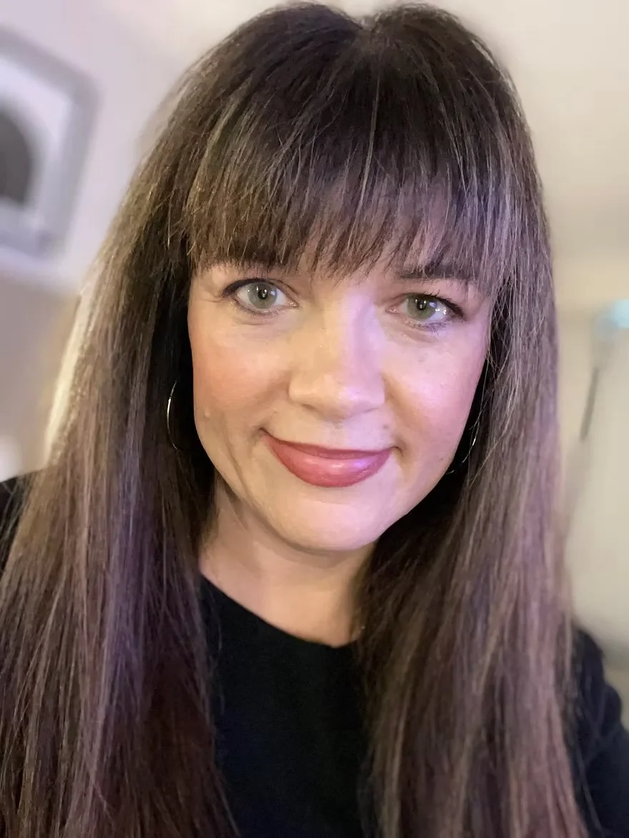 A woman with long brown hair and bangs, wearing hoop earrings, smiling at the camera.