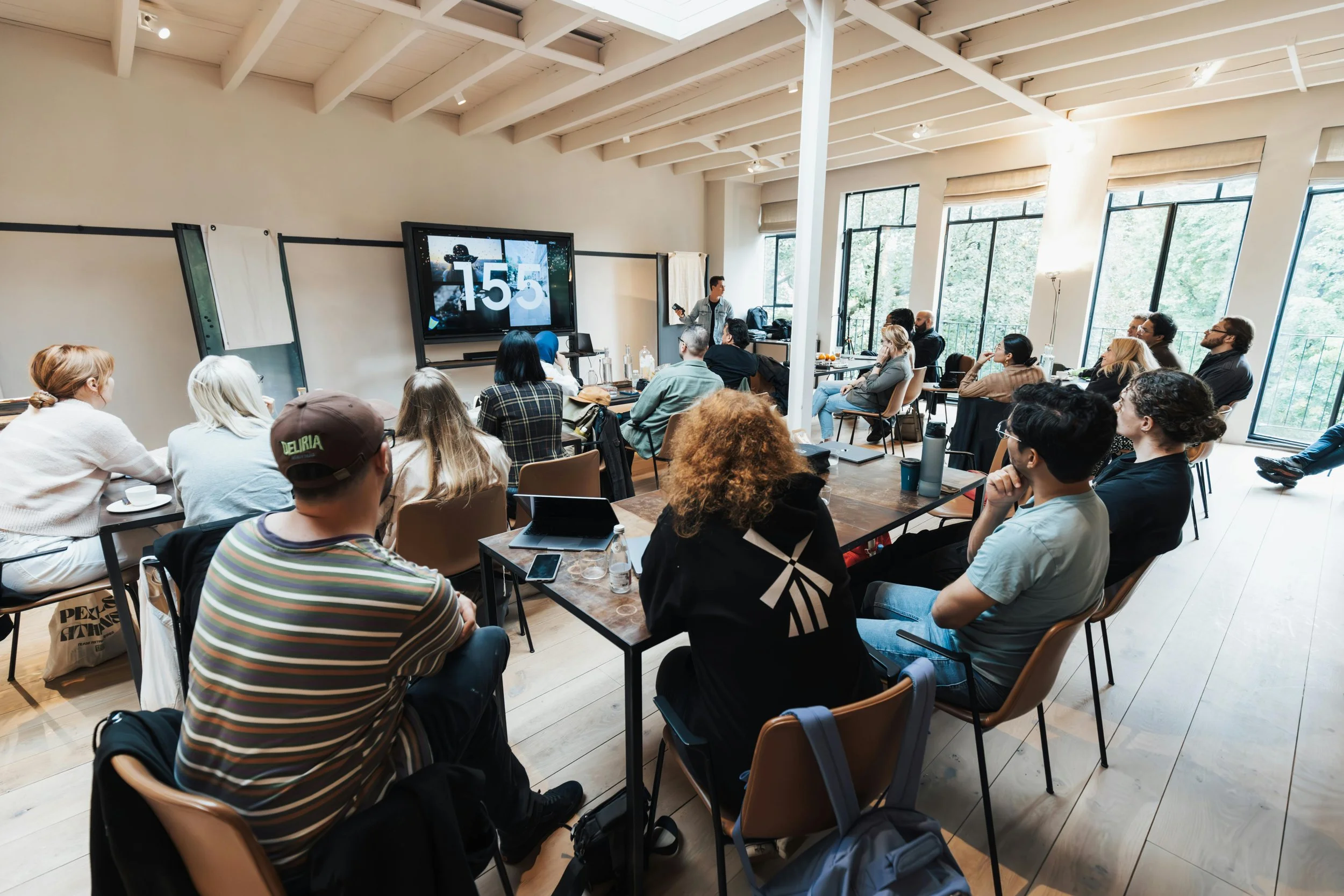 A group of people attending a workshop or presentation in a spacious room with large windows, some are watching a speaker at the front, and there is a large screen displaying the number '155'.