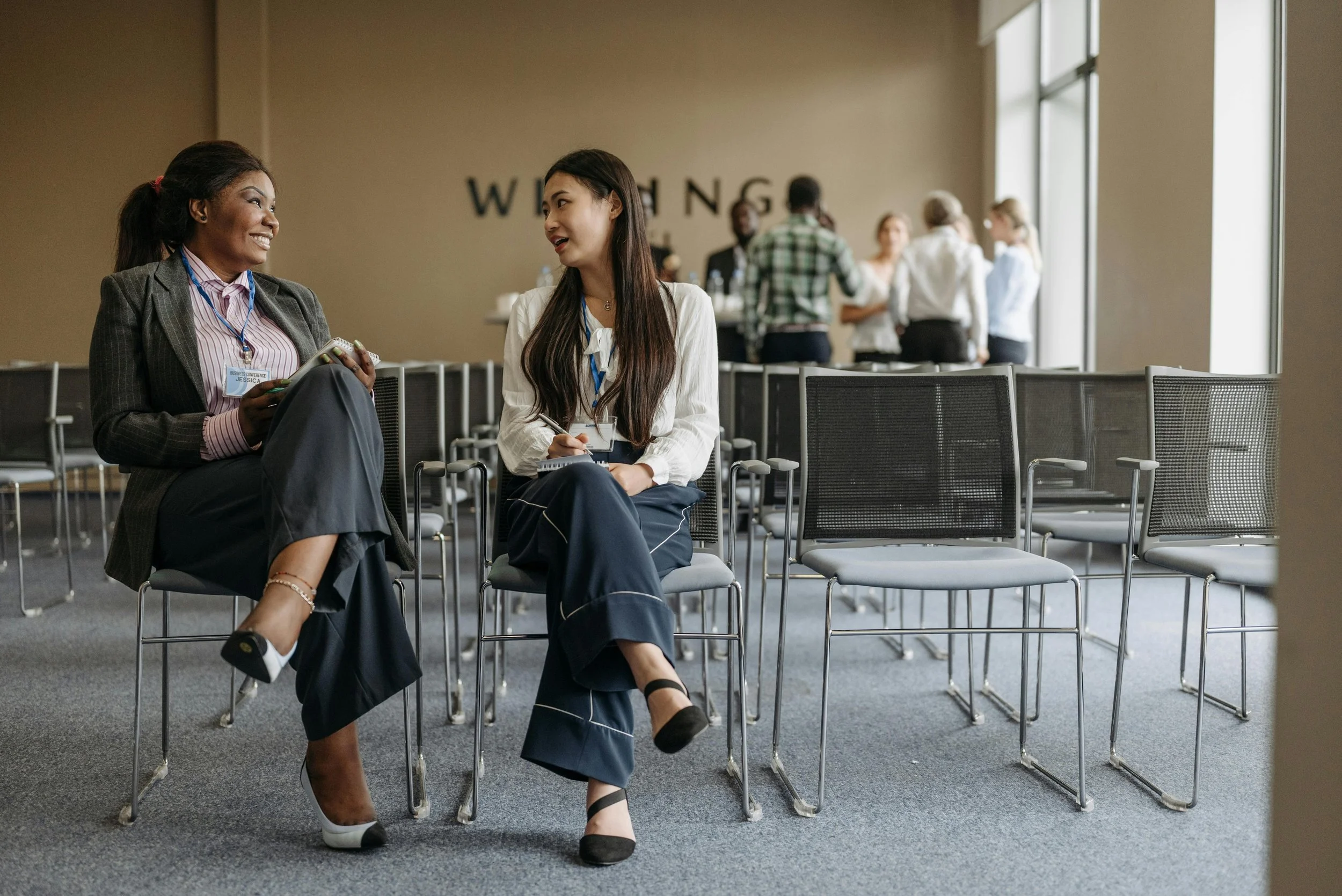 Two women are sitting and talking in a conference room with empty chairs around them. They are smiling and wearing business attire, with other people in the background having a discussion.