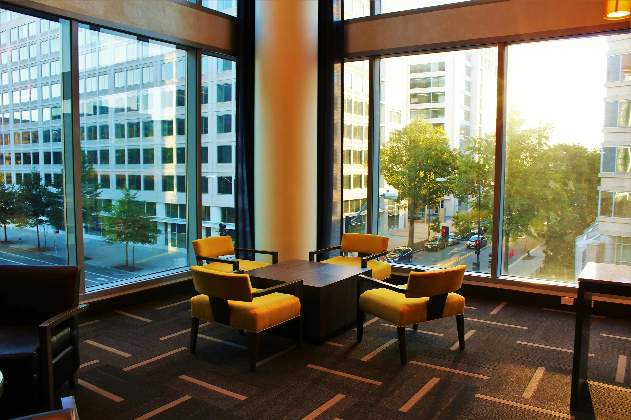 Interior of a modern office space with large floor-to-ceiling windows showing a city street with buildings, trees, and cars, and sunlight streaming inside. There are four yellow upholstered chairs around a small wooden table on a patterned dark carpet.
