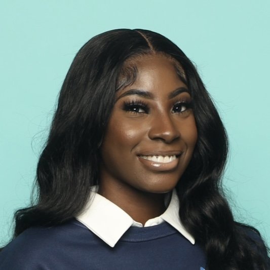 Portrait of a young black woman with long dark hair wearing a white collared shirt and a navy blue sweater, smiling against a light turquoise background.