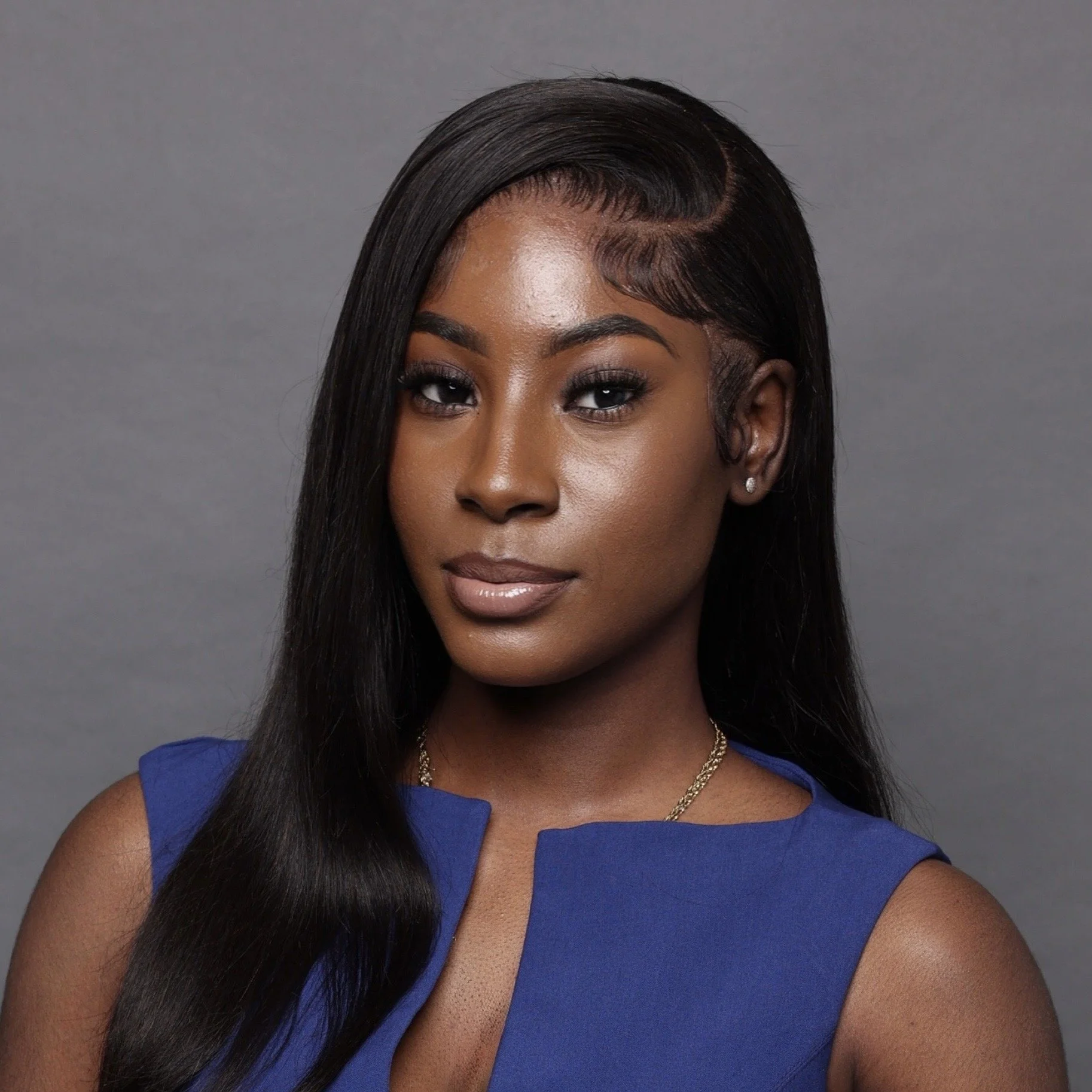 Portrait of a young African American woman with long black hair, wearing a blue sleeveless top, silver earrings, and a gold necklace, against a gray background.