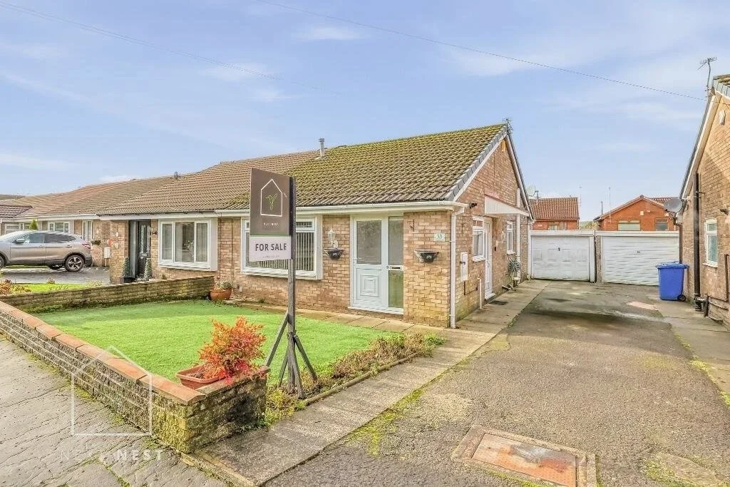 A brick house with a small front lawn and a "For Sale" sign in the yard. The house has a white door and windows, with a driveway leading to a white double garage door. A gray SUV is parked in front, and the sky is clear.