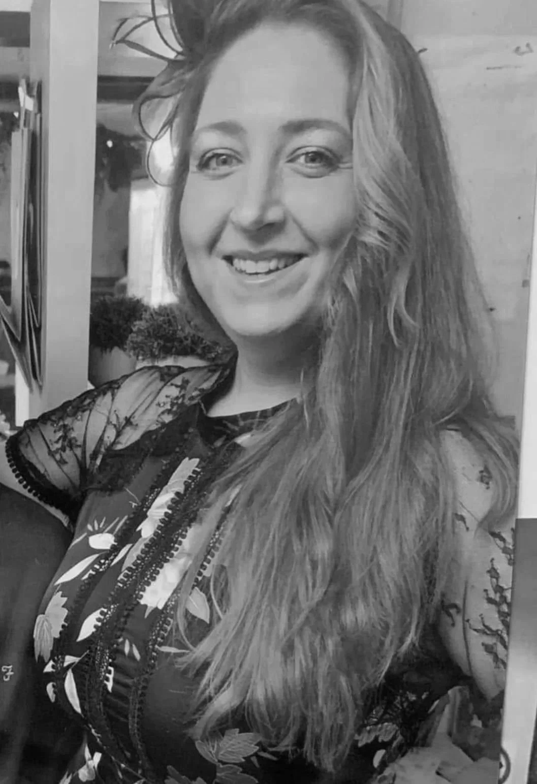 A smiling woman with long, wavy hair, wearing a floral-patterned top with lace details, in black and white.