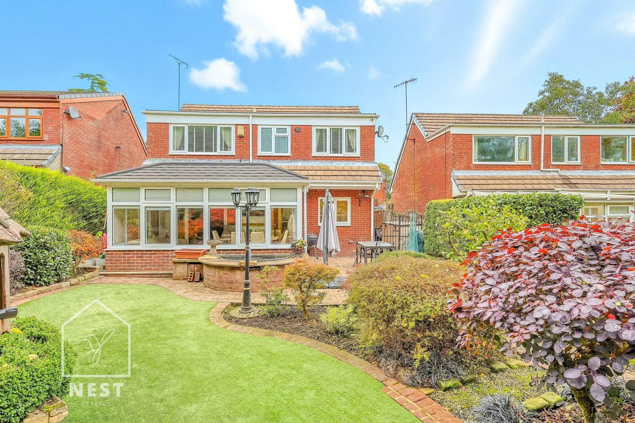 Backyard garden with a manicured lawn, bushes, trees, flowerbeds, brick patio, sunshade, patio table and chairs, lamppost, and a brick house with a conservatory and upper windows under a blue sky with scattered clouds.