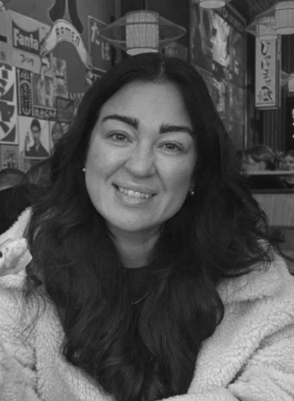 A woman with long dark hair, smiling in a restaurant with posters and signs on the wall behind her.