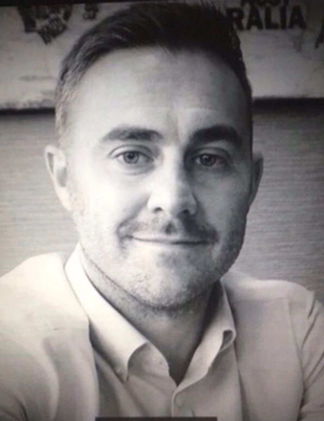 A black and white close-up portrait of a man with short hair, wearing a collared shirt, smiling slightly.