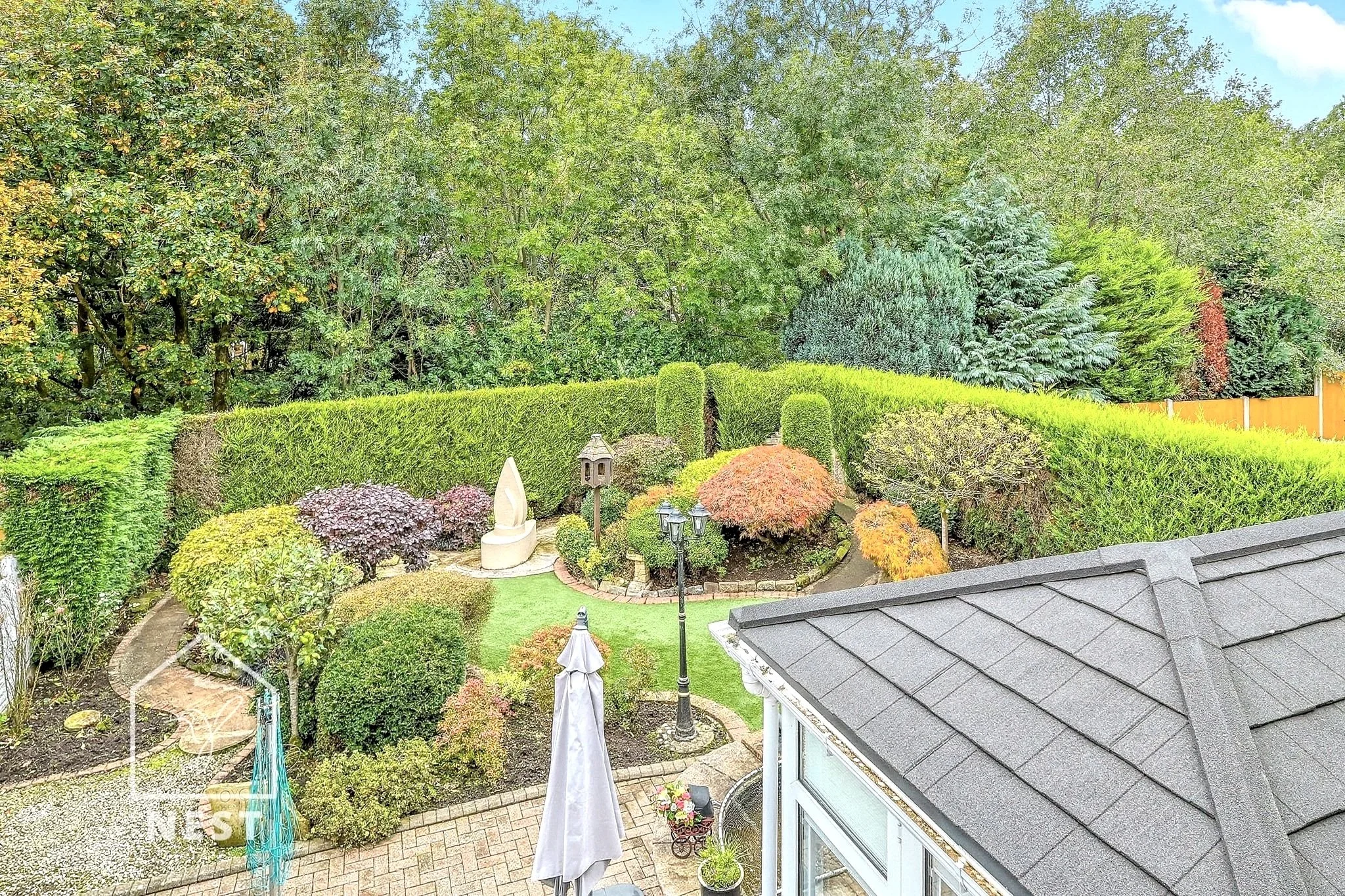 view of a backyard garden with neatly trimmed hedges, various bushes and trees, a stone pathway, a white outdoor sculpture, an umbrella, and a house roof in the foreground.