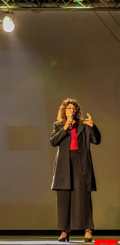 Laura Pasi con un tailleur nero pantalone e giacca a tre quarti, con in mano un microfono mentre gesticola