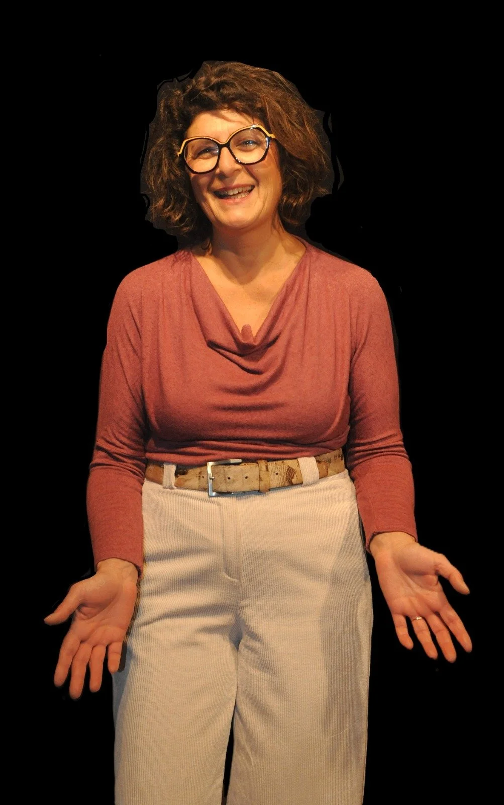 Laura Pasi, sorridente con occhiali grandi, capelli ricci e vestita con una maglietta color vinaccia, pantaloni e cintura color cintura beige, che fa un gesto con le mani.