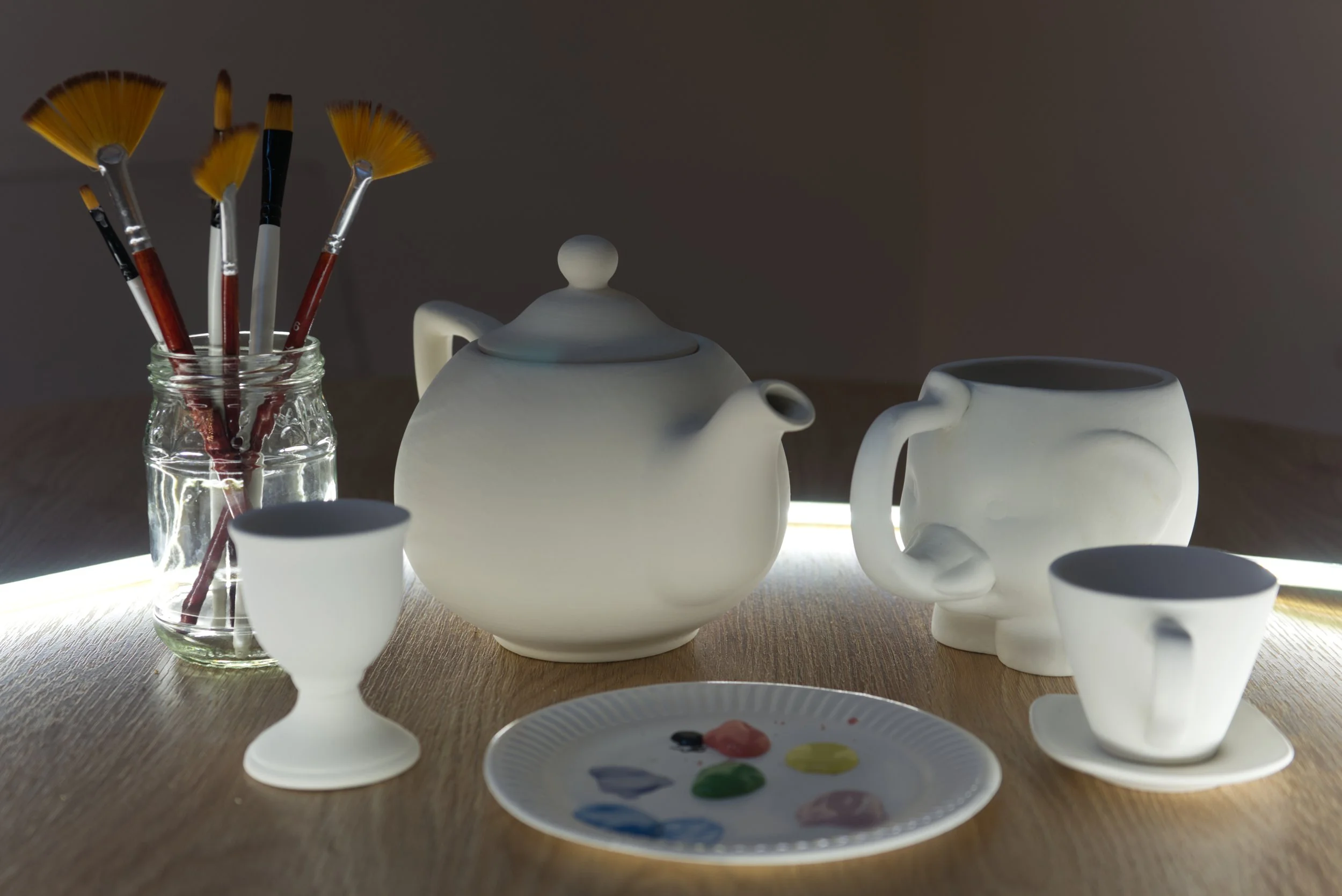 Keramiktassen, Teekanne, Pinsel in einem Glas, Malpalette mit Farben auf einem Holztablett, auf einem Tisch.