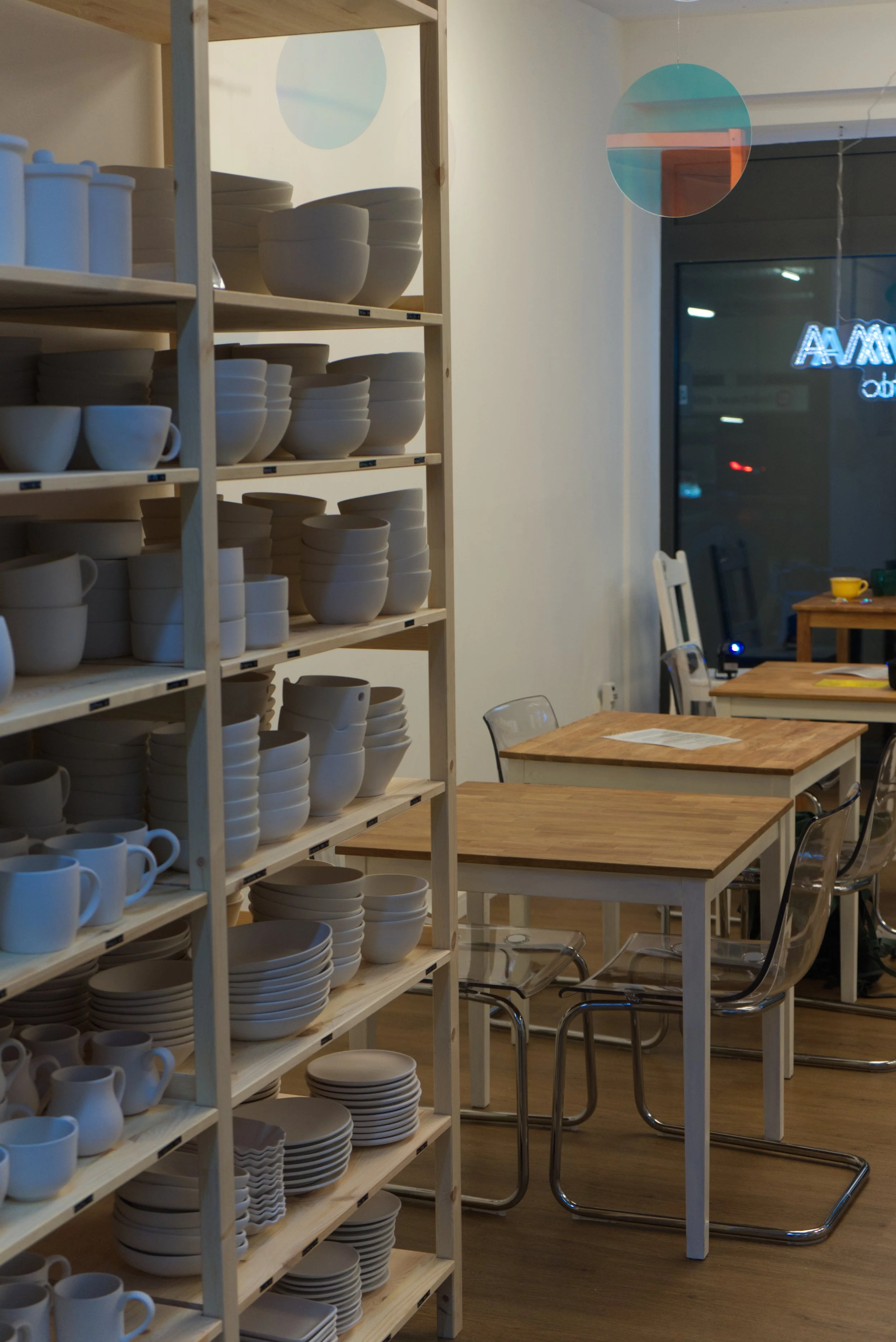 Regal mit weißen Geschirr in einem Ladenlokal, mit Tischen und Stühlen im Hintergrund, vor einem Fenster mit Neonbeleuchtung im Café-Stil.