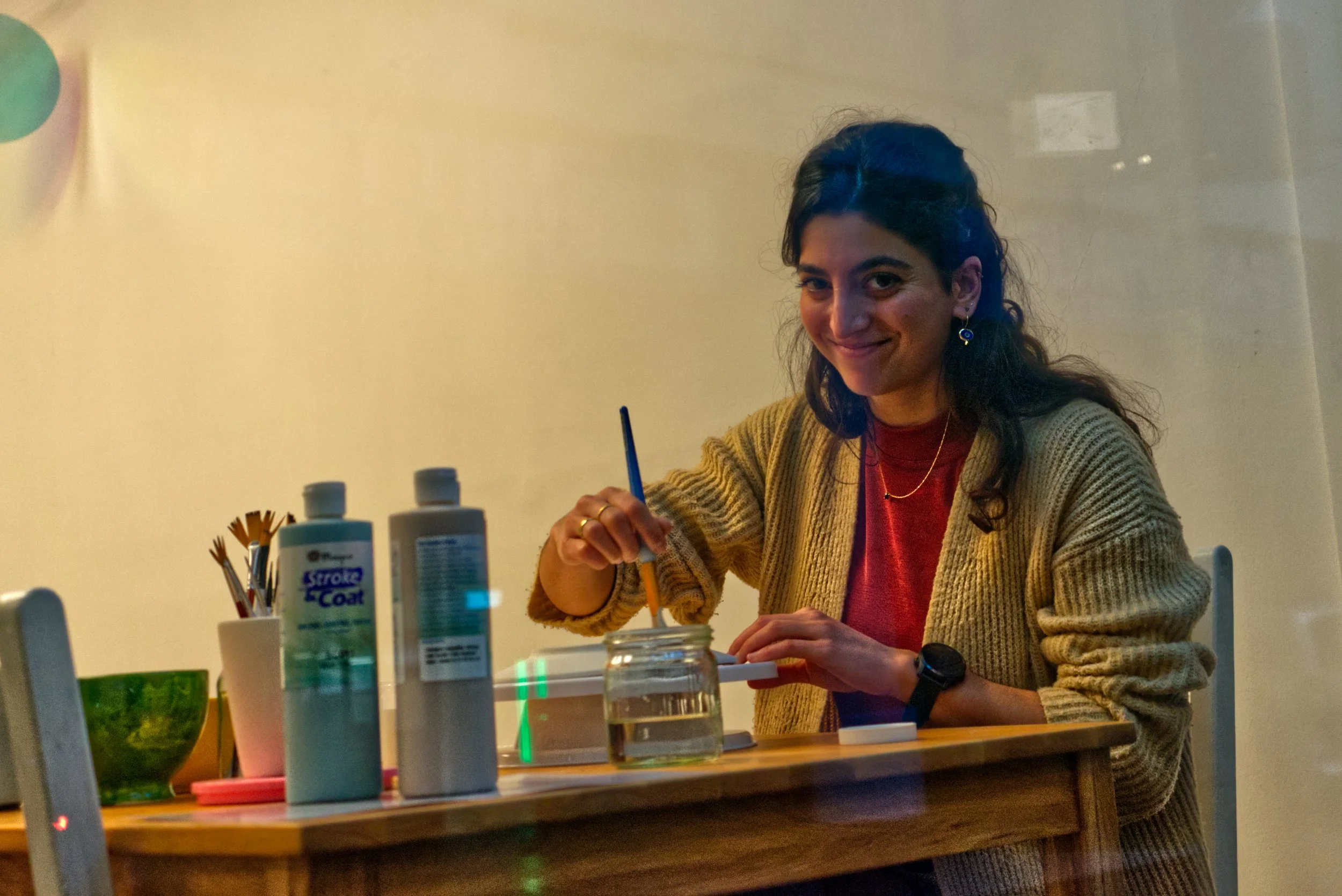 Eine Frau sitzt an einem Tisch, hält einen Pinsel in der Hand, arbeitet an einem Kunstprojekt und lächelt. Auf dem Tisch stehen Gebäude, Körperpflegemittel, Pinsel und ein Glas Wasser.