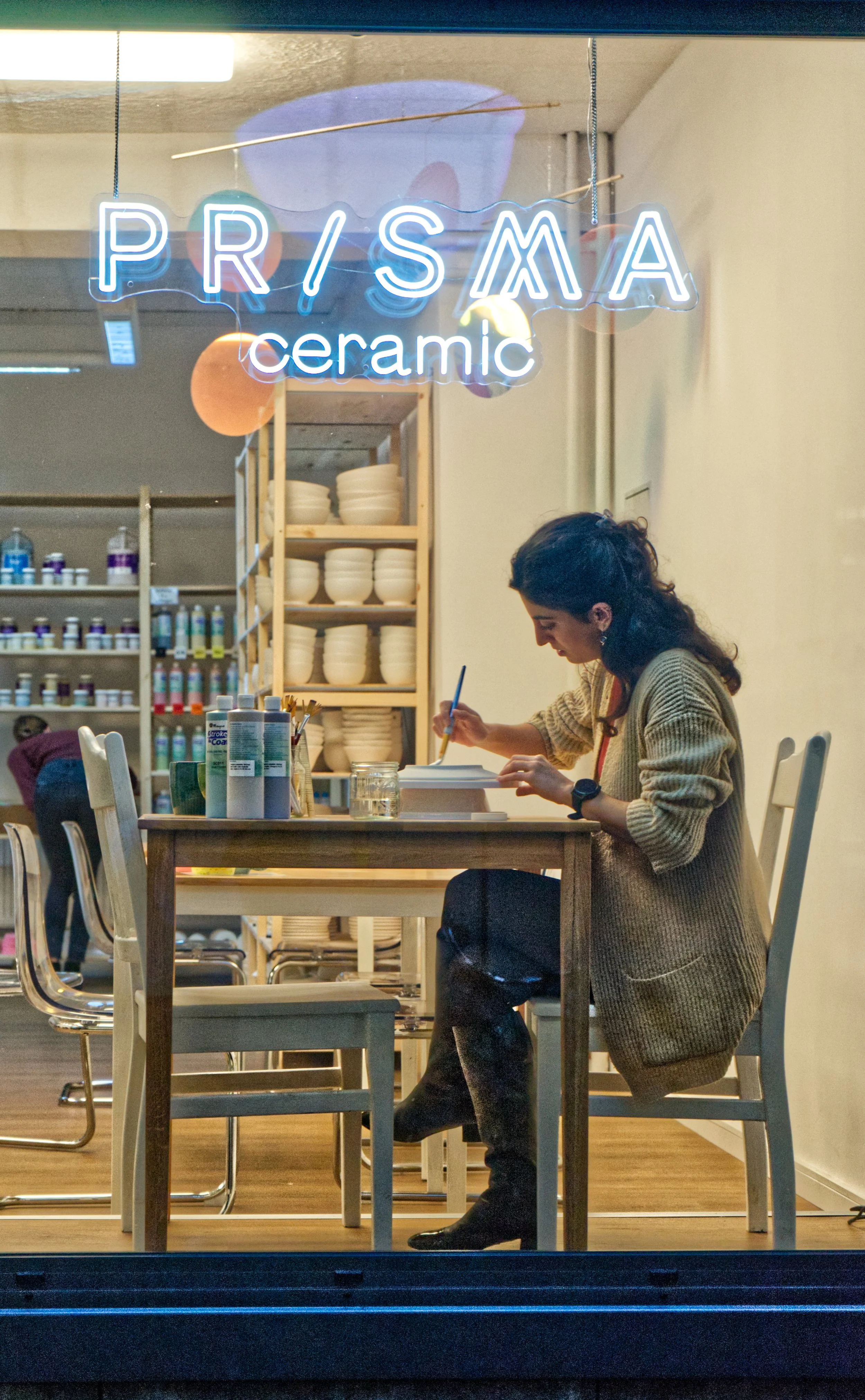 Eine Frau, die an einem Tisch in einem Keramikgeschäft arbeitet, bemalt oder graviert eine Keramikschüssel. Im Hintergrund sind Regale mit weiteren Keramikartikeln und Farben. Eine Neonreklame hängt im Fenster mit der Aufschrift 'PRISMA ceramic'.