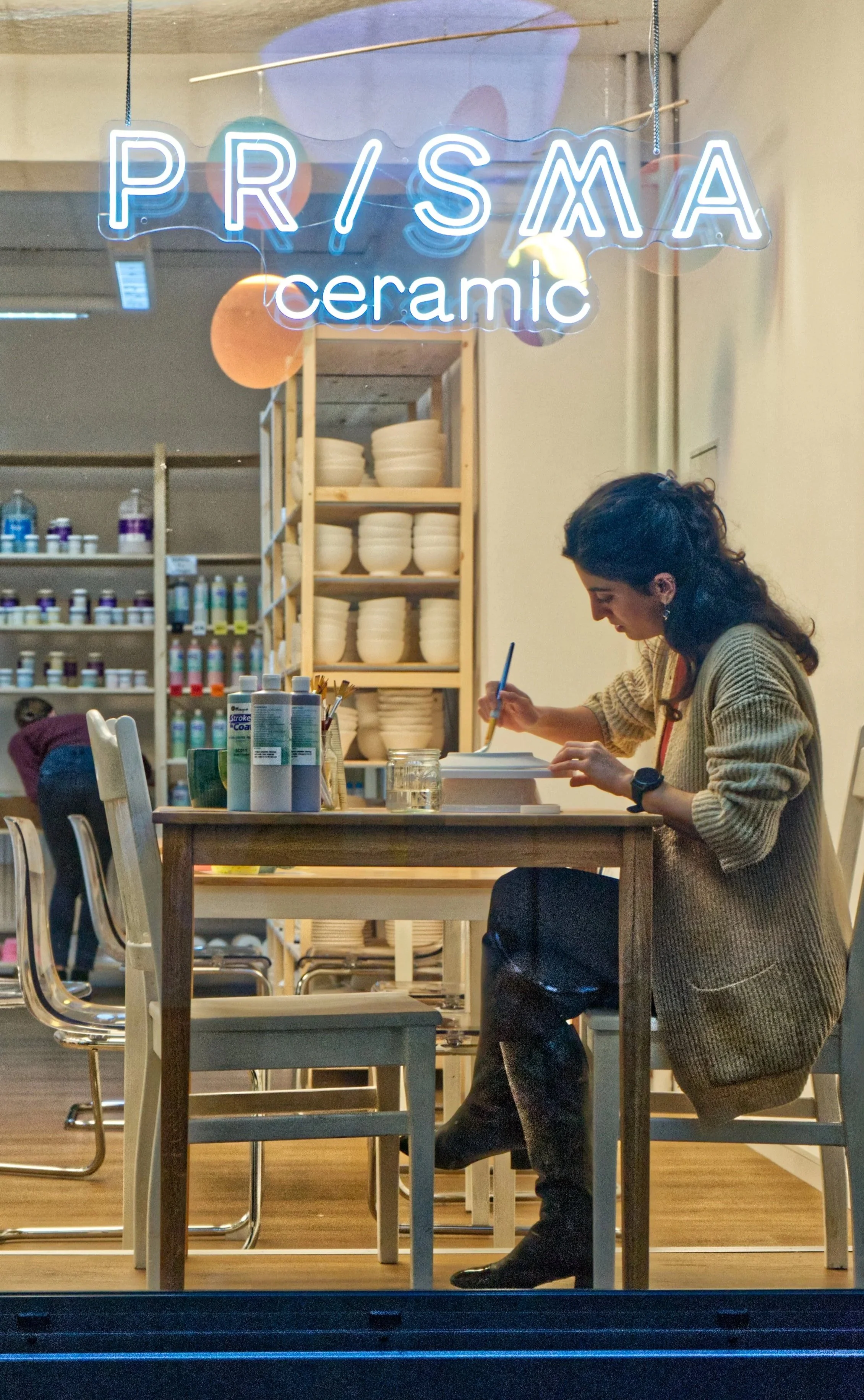 Eine Frau sitzt an einem Holztisch in einem Geschäft, schreibt oder zeichnet in ein Buch, im Hintergrund sind Regalregale mit Keramiktellern und Farben ausgestellt, die Geschäftsschriftzug ist in Neonlicht geschrieben und lautet "PRISMA ceramic".