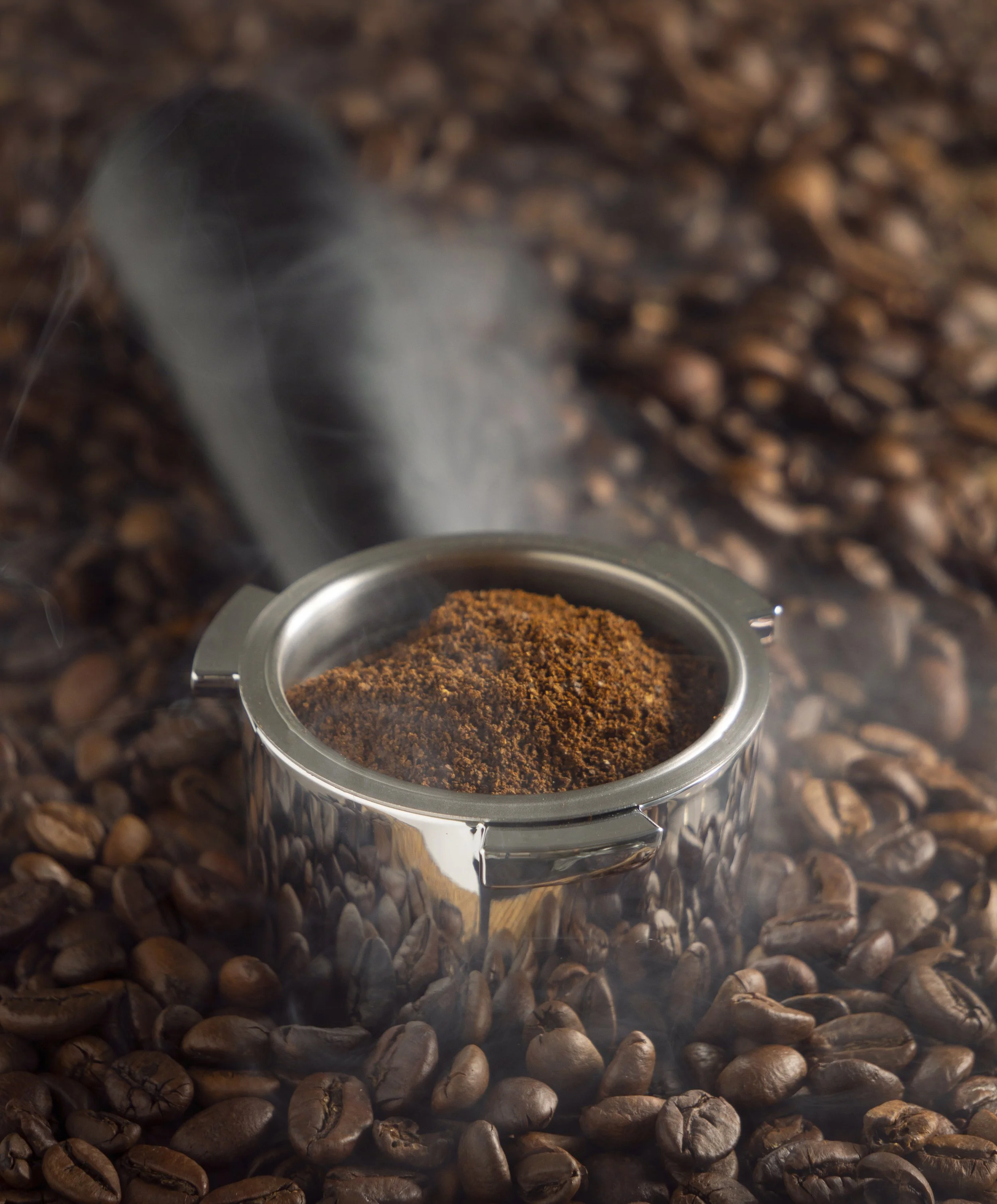 Close-up of a metal container filled with ground coffee on a bed of roasted coffee beans with steam rising.