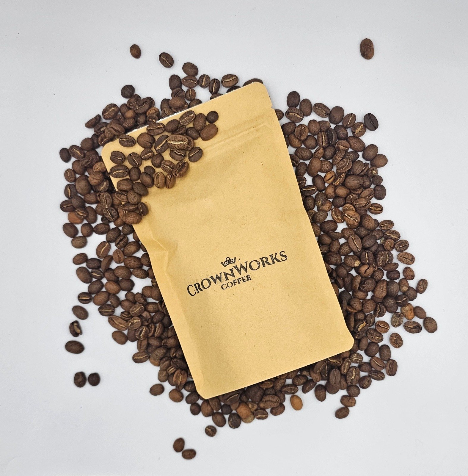 A pile of roasted coffee beans with a yellow paper packet labeled 'CROWNWORKS COFFEE' placed on top, on a white surface.