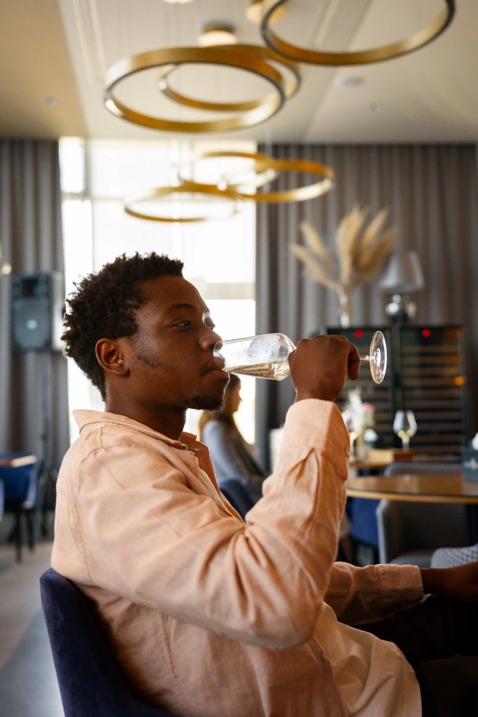 A young man with curly hair in a beige shirt sitting in a restaurant, drinking white wine from a glass.