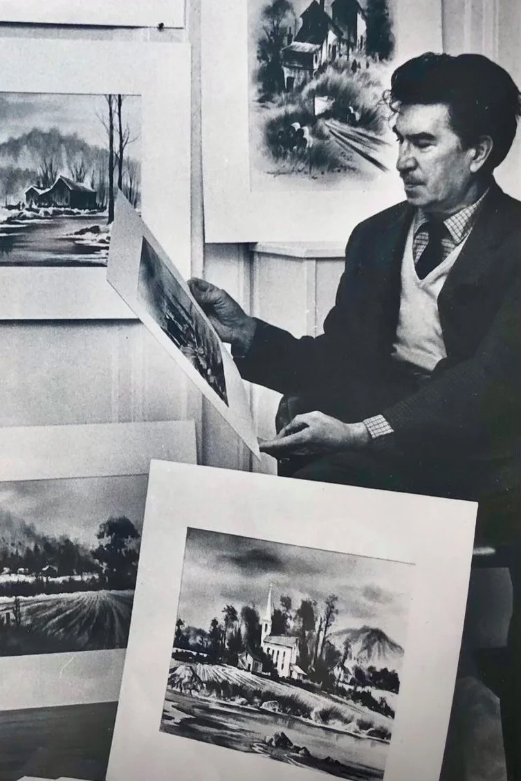 A man in a blazer and sweater vest examining paintings of landscapes at an art exhibition, with several landscape artworks displayed on the walls and table.