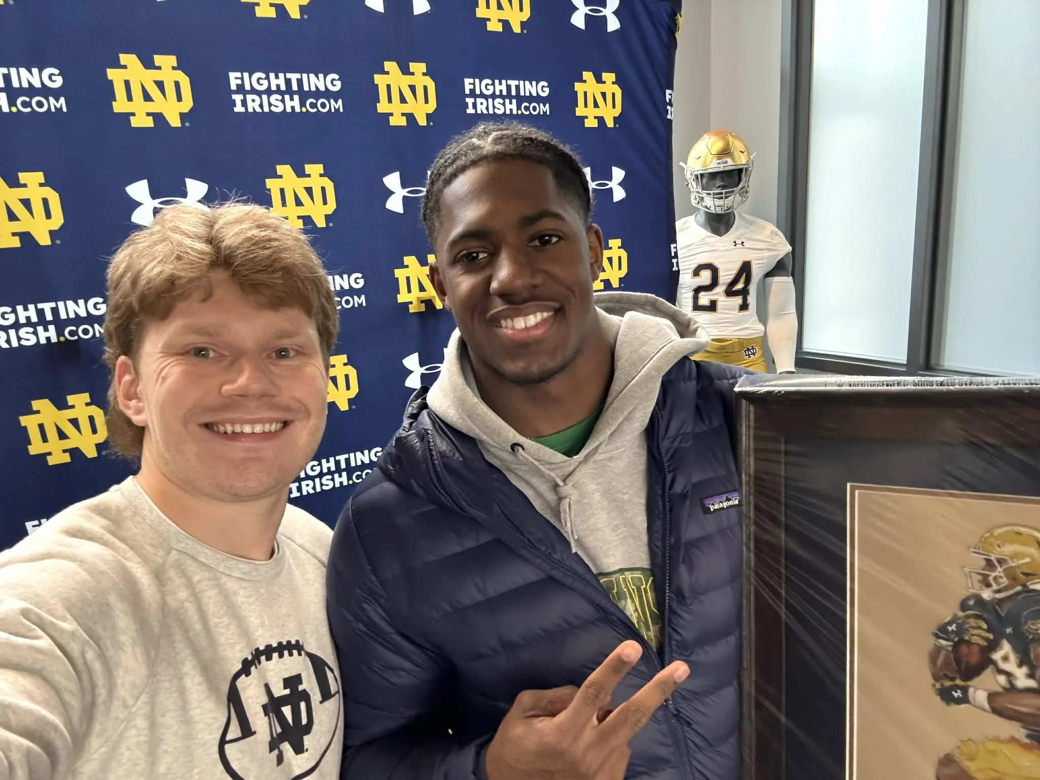 Two smiling men taking a selfie at a Notre Dame Fighting Irish event, with Notre Dame logos and a mannequin in football gear in the background.