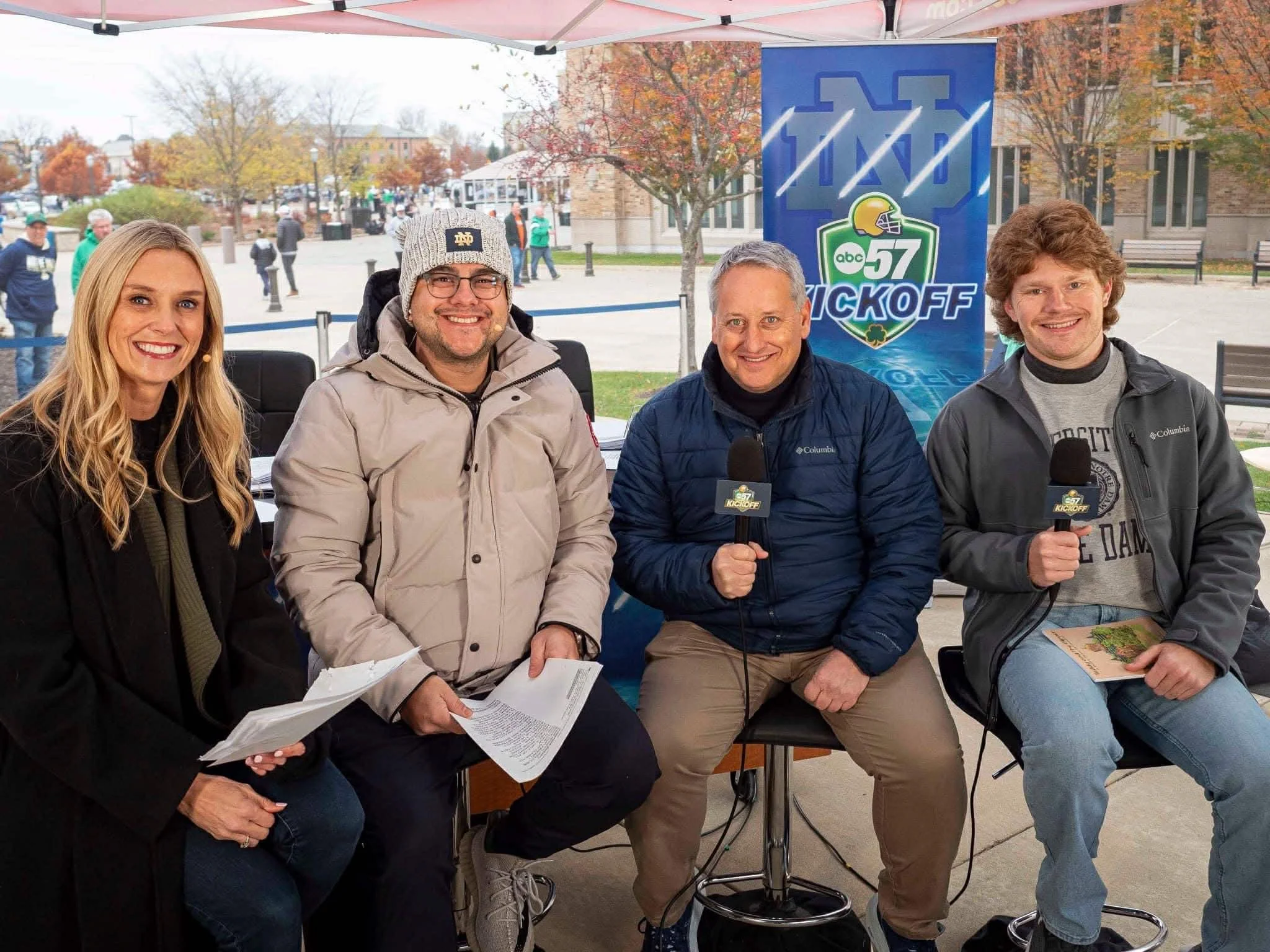 Four people sitting at a broadcast table outdoors with microphones, a banner behind them reading 'ABC 57 Kickoff,' and people walking in the background.