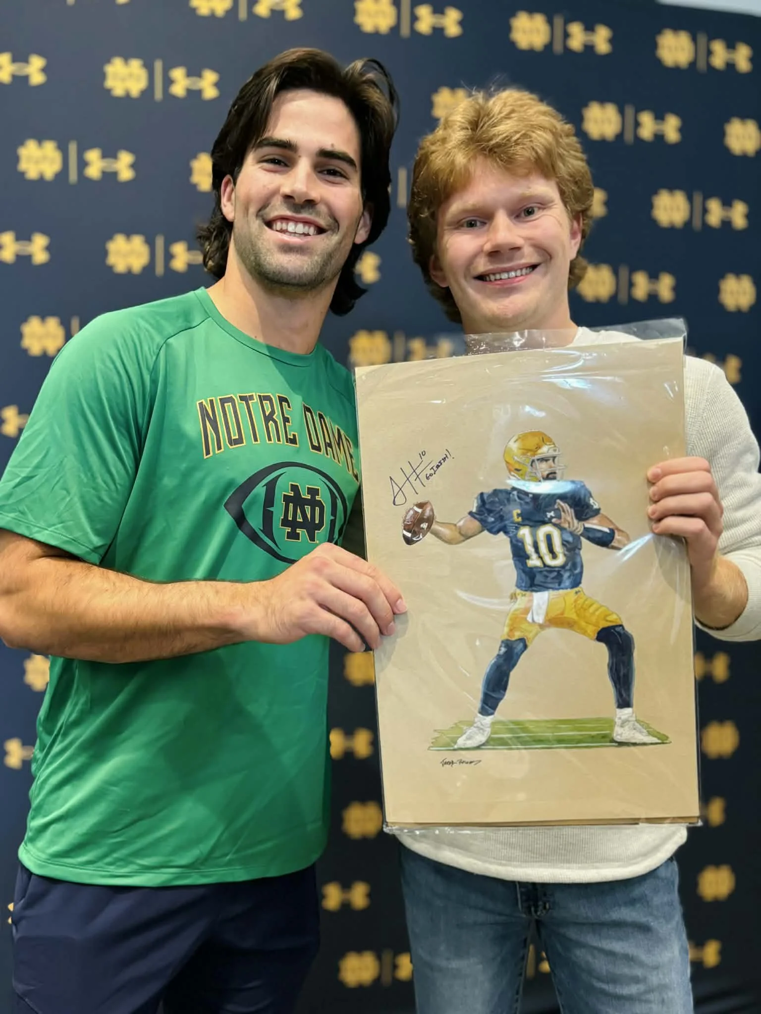 Two smiling young men standing in front of a backdrop with yellow Under Armour logos, holding a framed artwork of a football player in a Notre Dame uniform with a $10 bill on the artwork's shoulder. One man is wearing a green Notre Dame shirt and the other a white sweater. The man in the white sweater is also holding a signed print of the artwork.