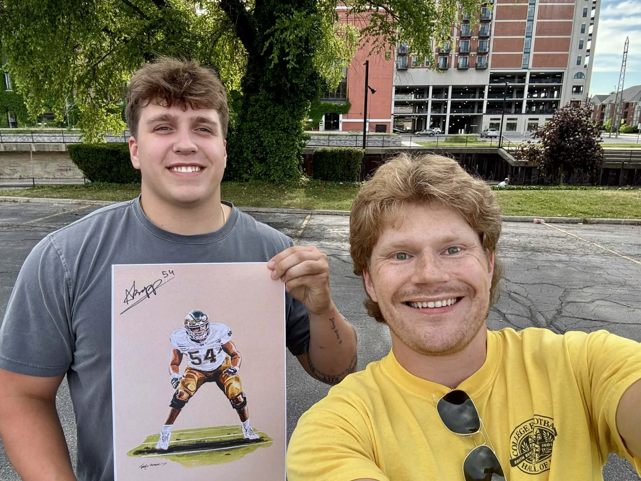 Two young men taking a selfie outdoors, one holding a signed poster of a football player with the number 54. Behind them is a parking lot, green trees, and a multi-story building.