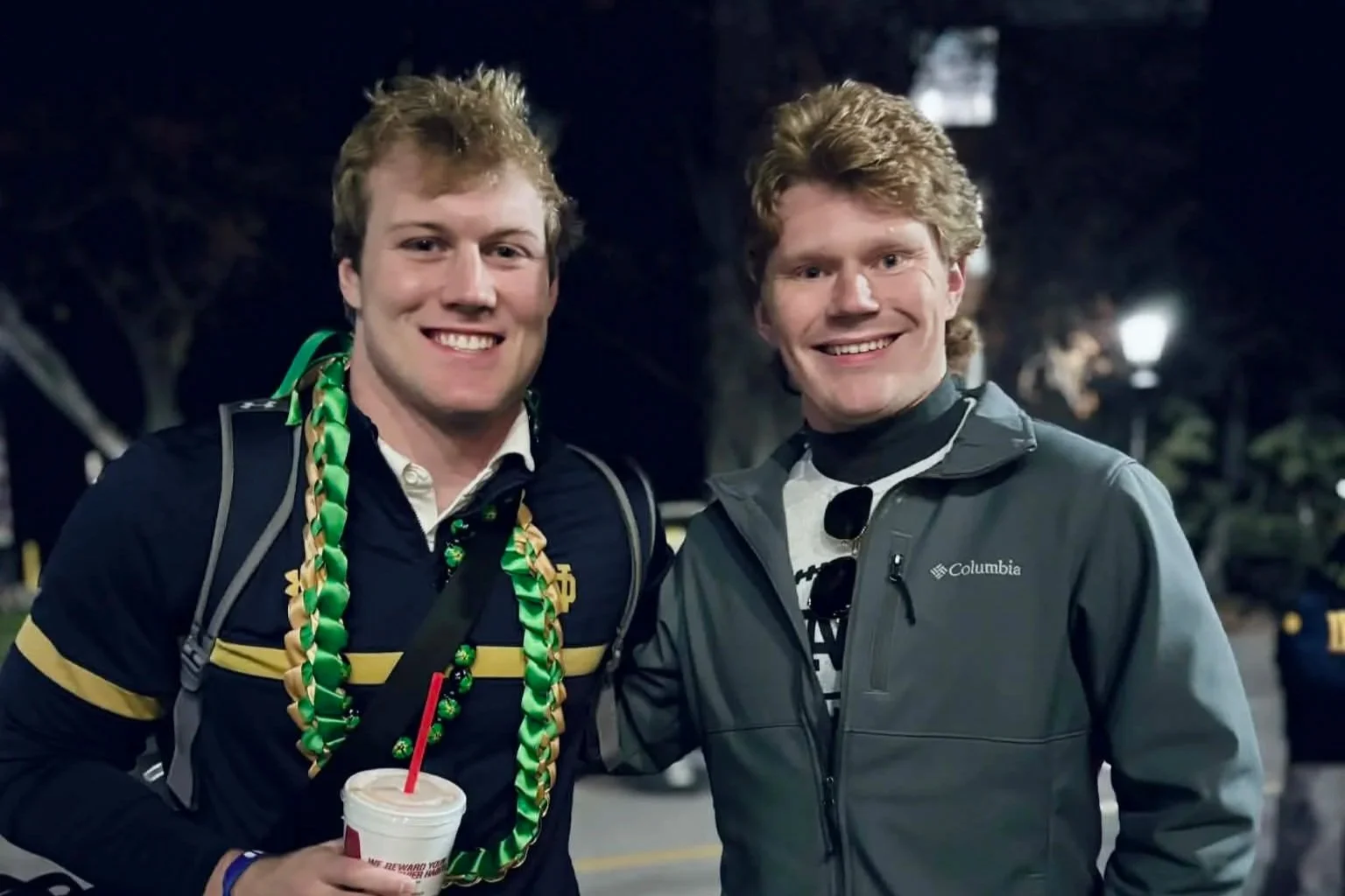 Two young men smiling at night outdoors, one holding a drink, the other wearing sunglasses, with streetlights and trees in the background.