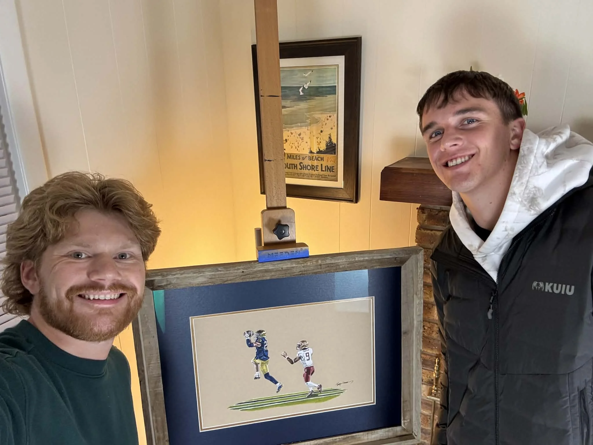 Two smiling men taking a selfie next to a framed artwork of two football players in action, in a cozy room with a wooden beam and wall decorations.