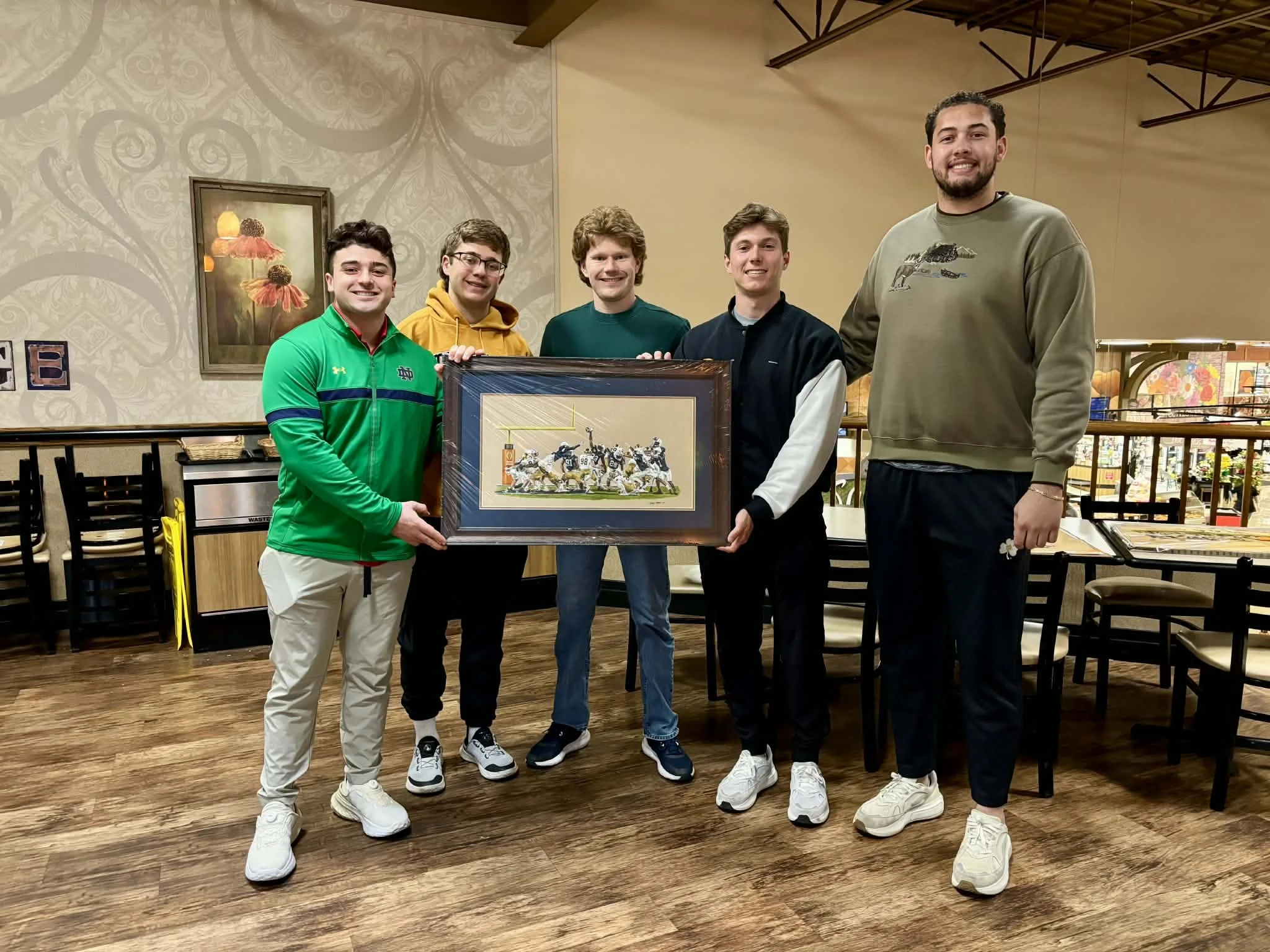 Five young men standing indoors, holding a framed artwork depicting the 2025 orange bowl, smiling at the camera.