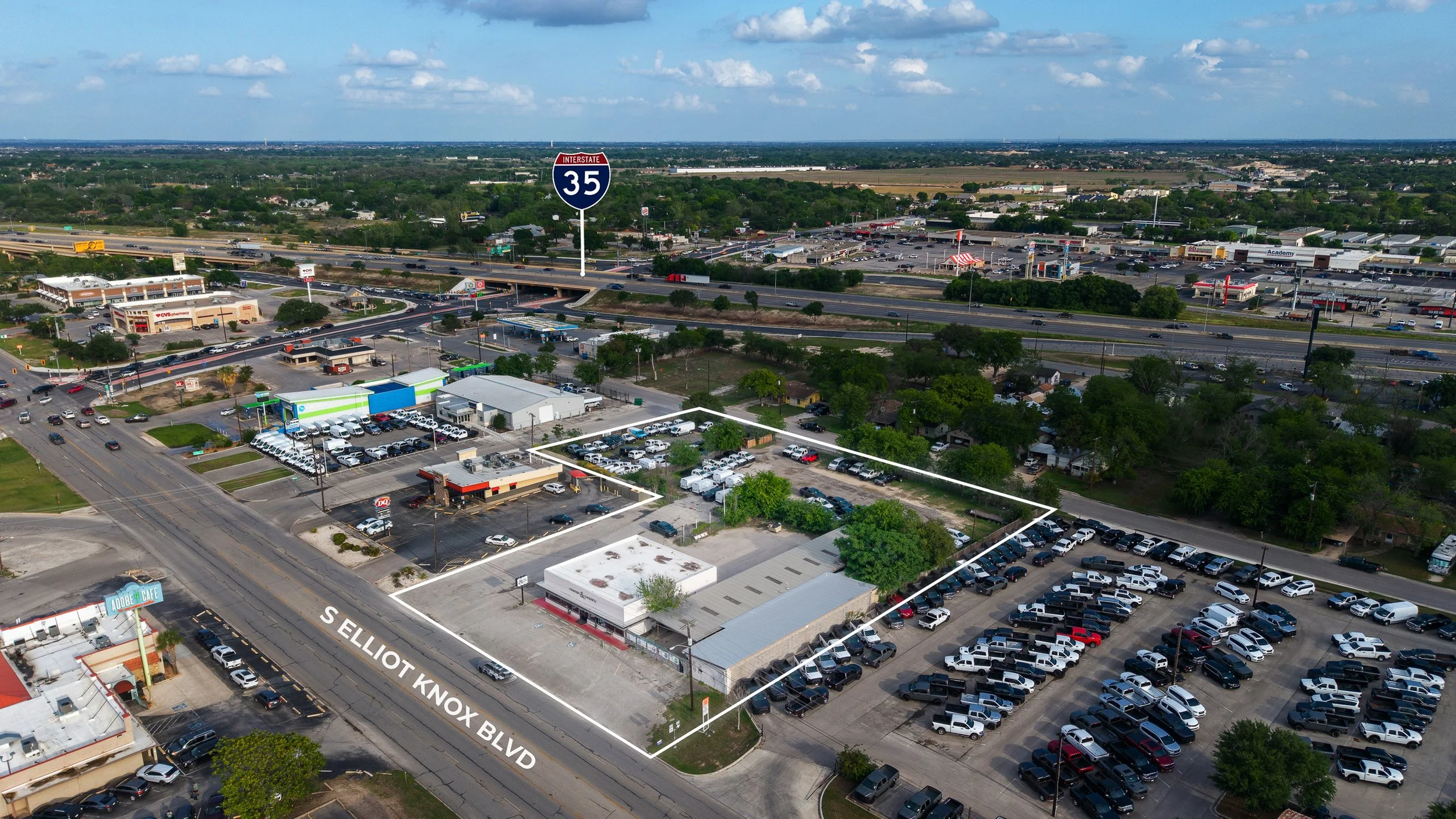 Aerial view of a busy commercial area with parking lots, stores, a highway, and green trees under a partly cloudy sky. The streets are labeled S Elliott Knox Blvd and the image features multiple cars, businesses, and a billboard.