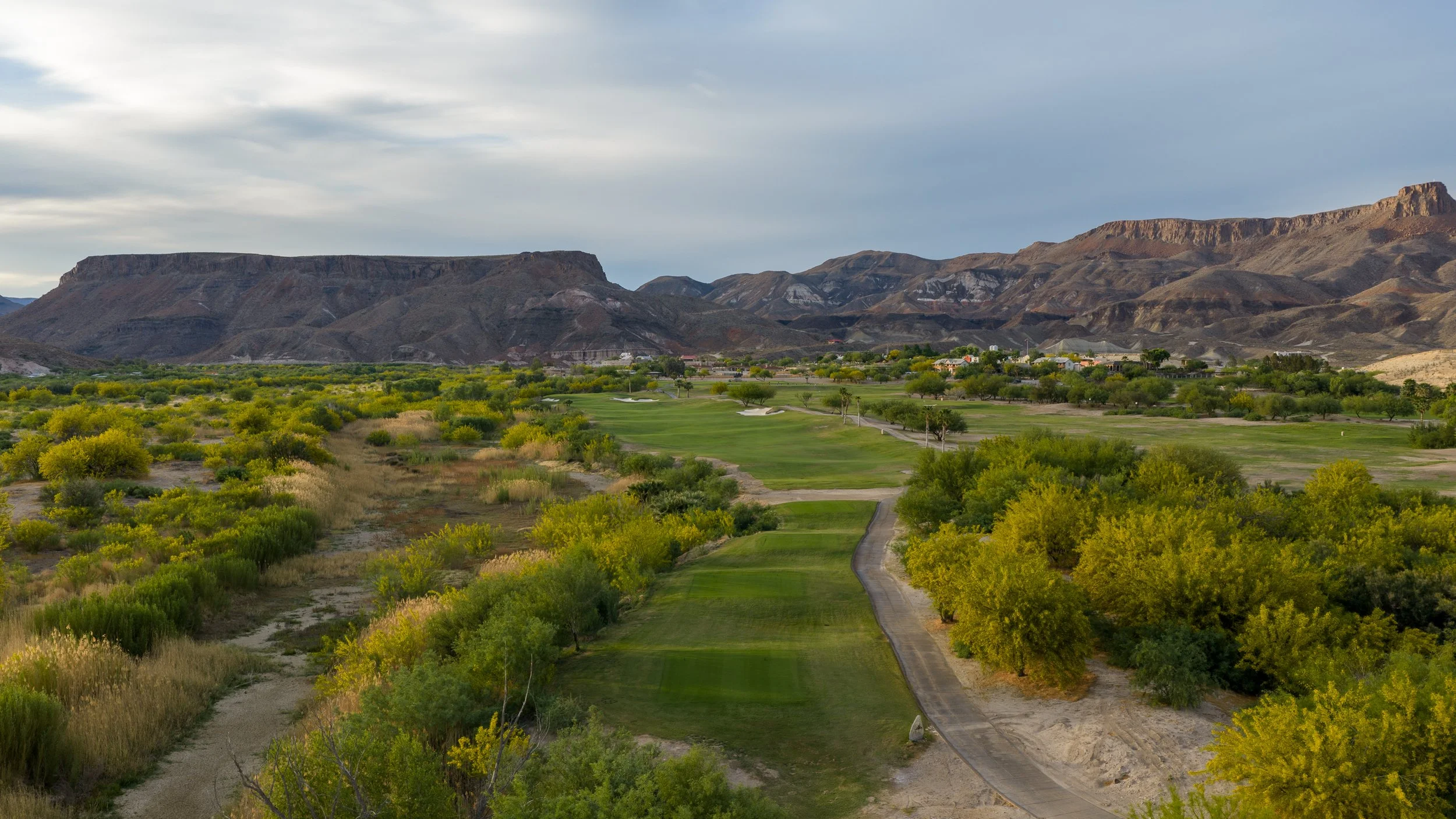 A golf course with green fairways, surrounded by trees and bushes, set against a backdrop of rocky, brown mountains and a cloudy sky.