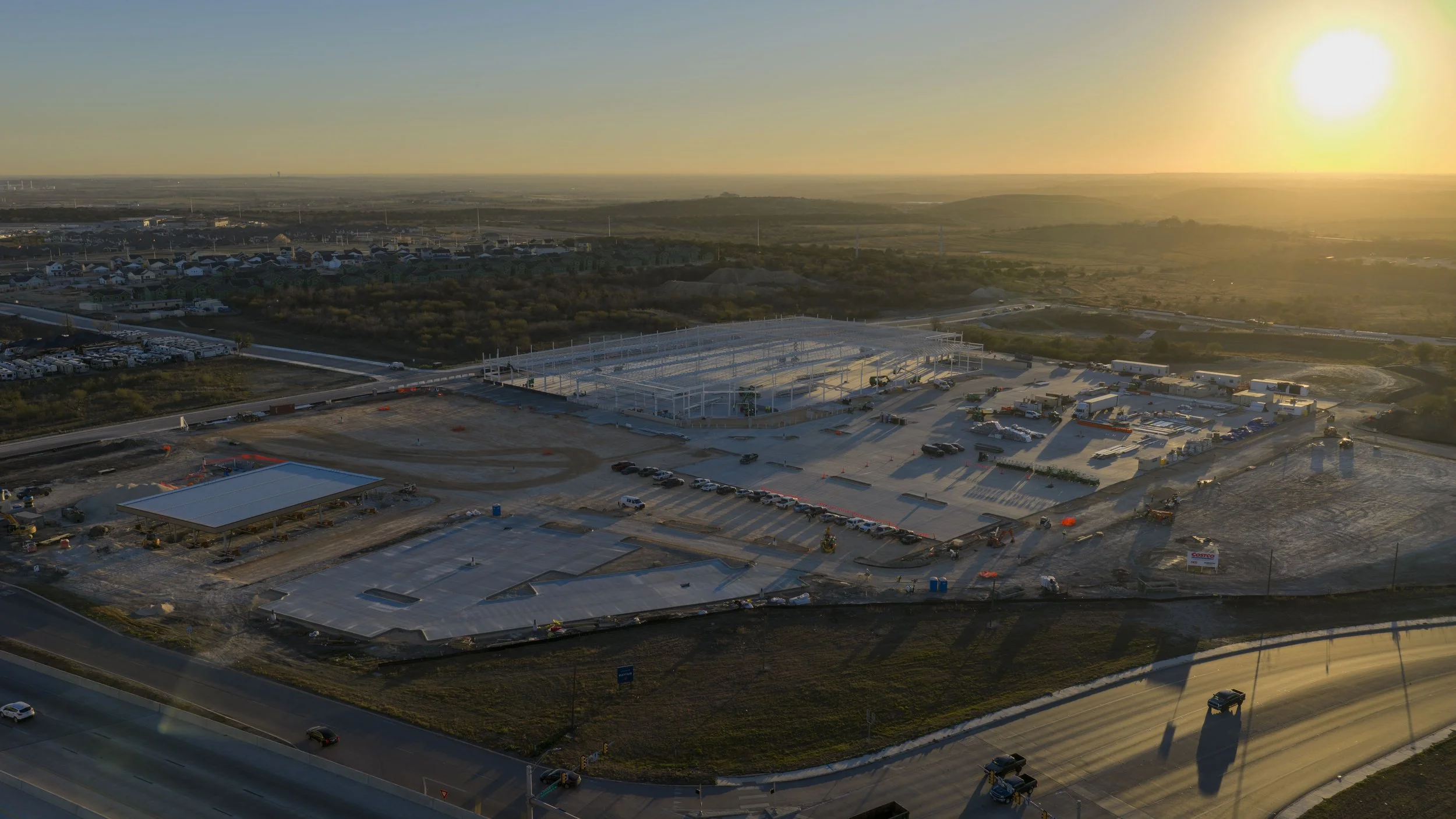 Aerial view of a construction site and nearby residential area at sunset with an open framework structure, vehicles, and equipment.
