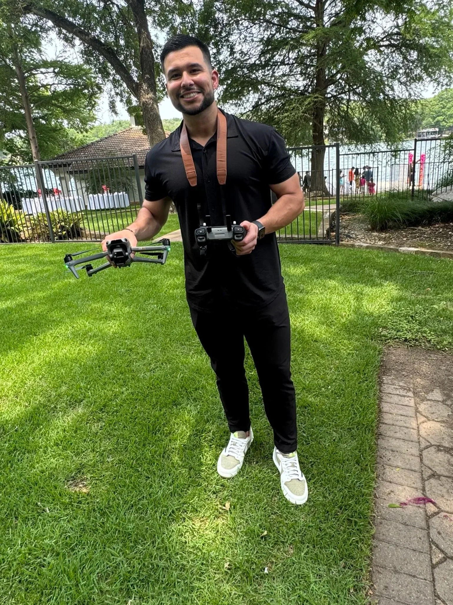 A smiling man in a black polo shirt and black pants holding a drone and its controller in a grassy outdoor area with trees, a water body, and a fence in the background.