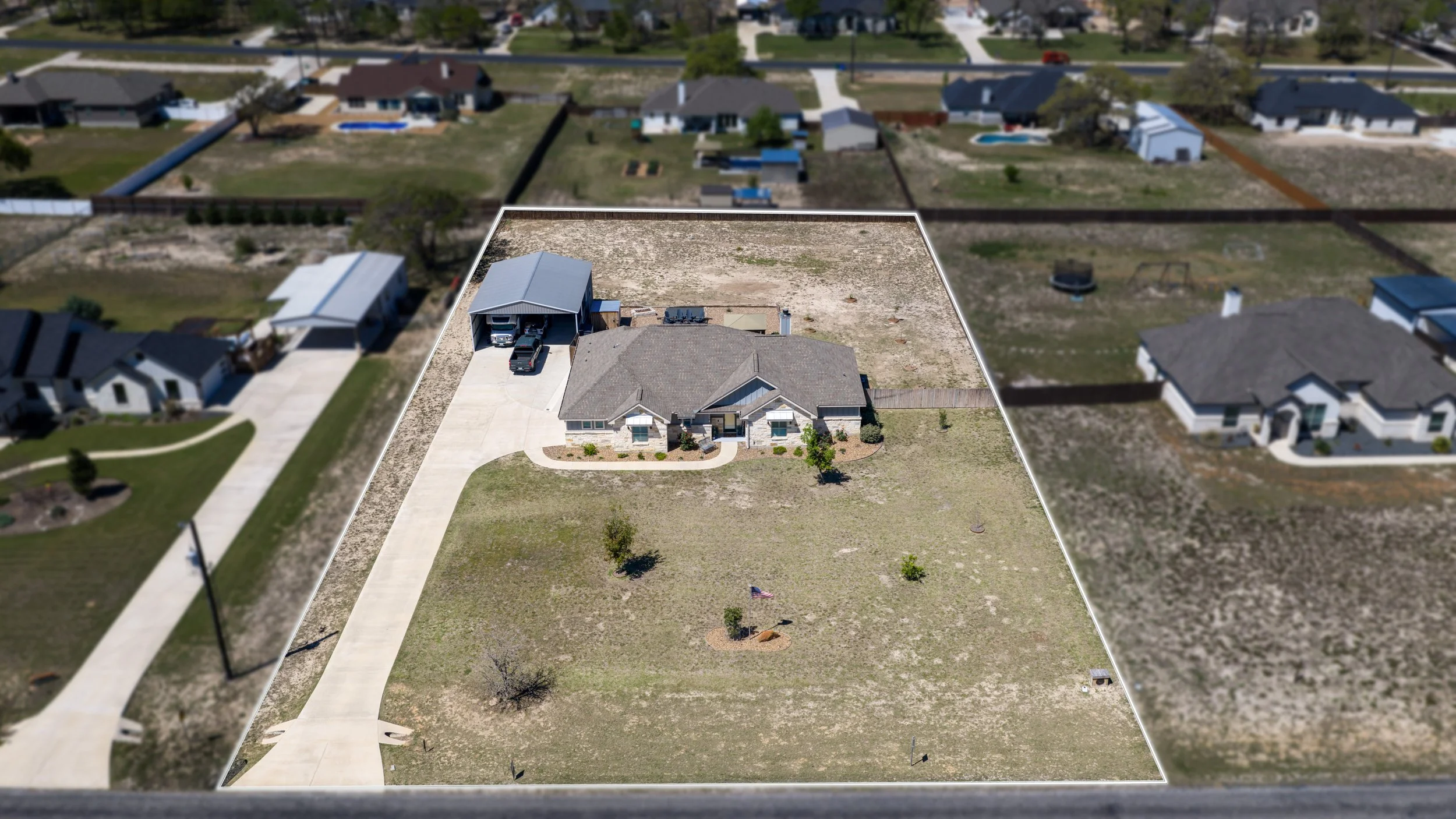 A single-family house with a front yard, driveway, and a detached garage on a large plot of land in a suburban neighborhood. The backyard is spacious and partly fenced, with a few small trees and sparse grass.