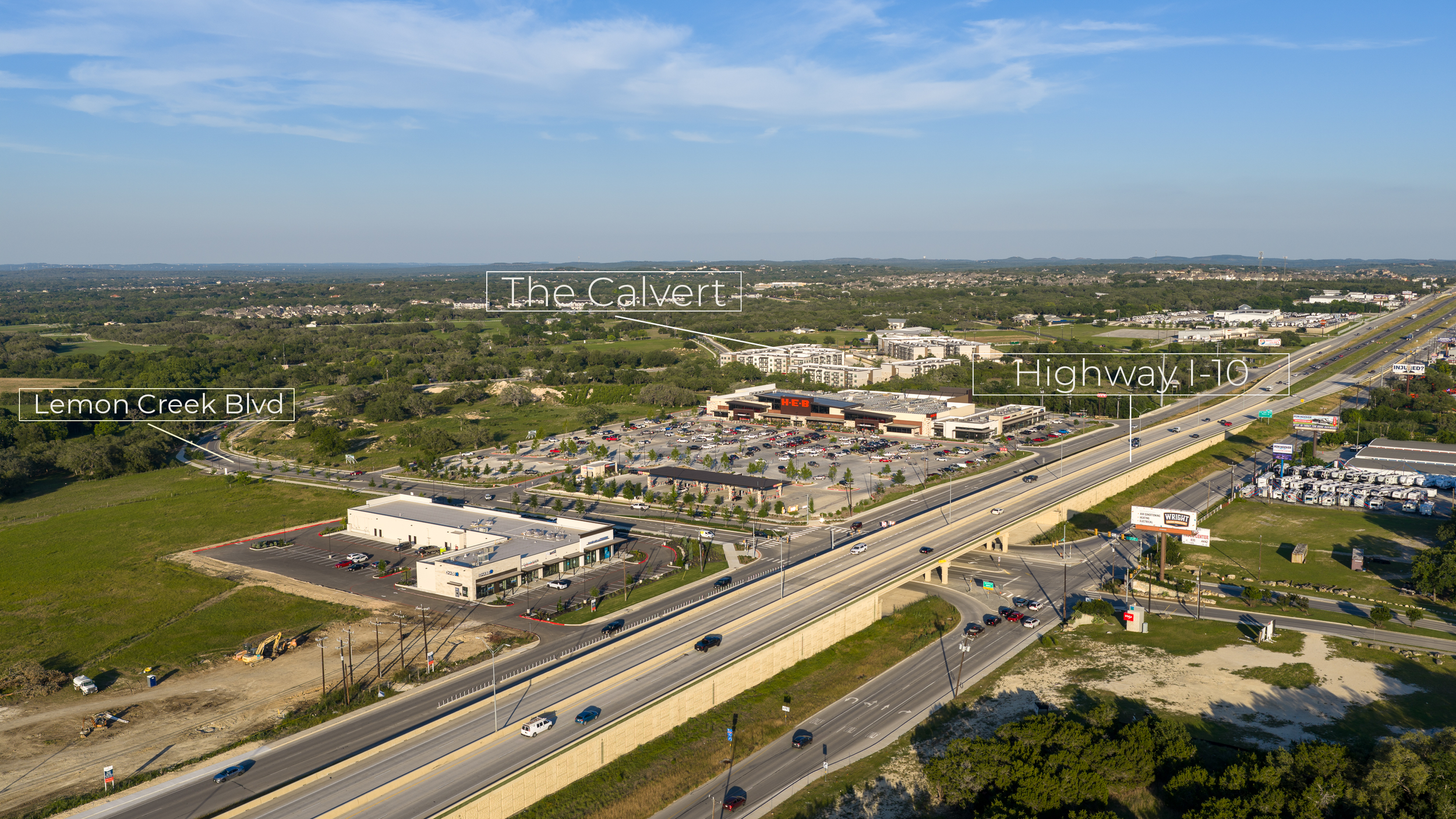 Aerial view of a commercial area with a shopping complex, busy highway, and surrounding greenery, labeled with street names and landmarks.