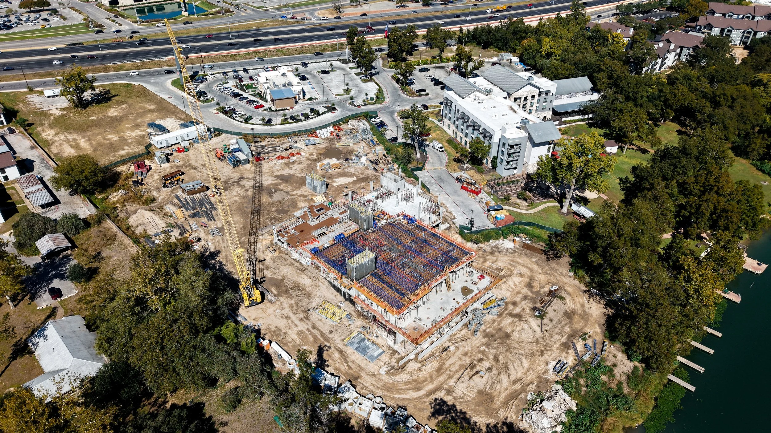 An aerial view of a construction site with a building foundation in progress near a body of water, surrounded by trees and nearby residential and commercial buildings.