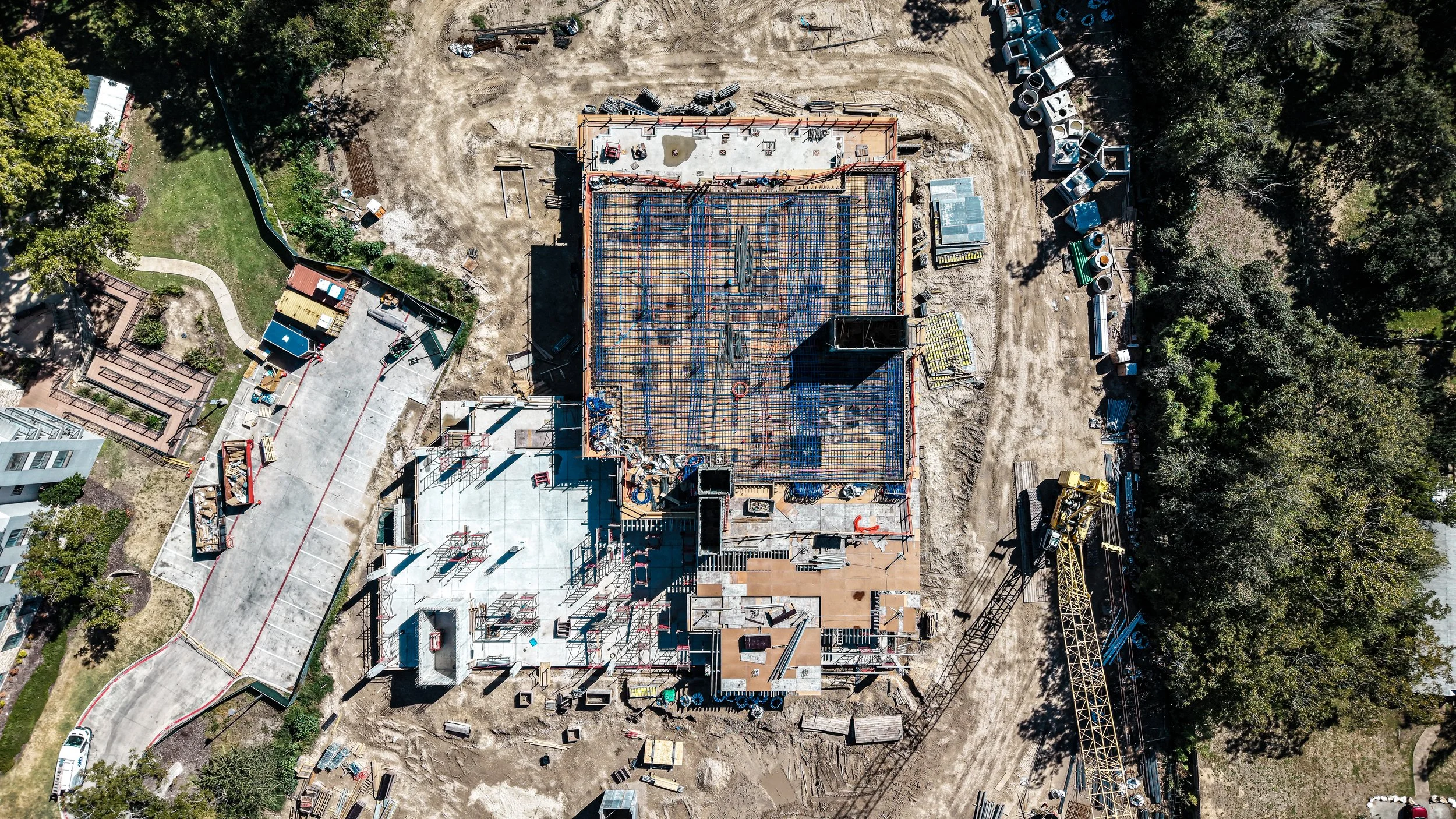 Aerial view of a construction site with a building frame, construction equipment, and materials, surrounded by trees and partially paved ground.