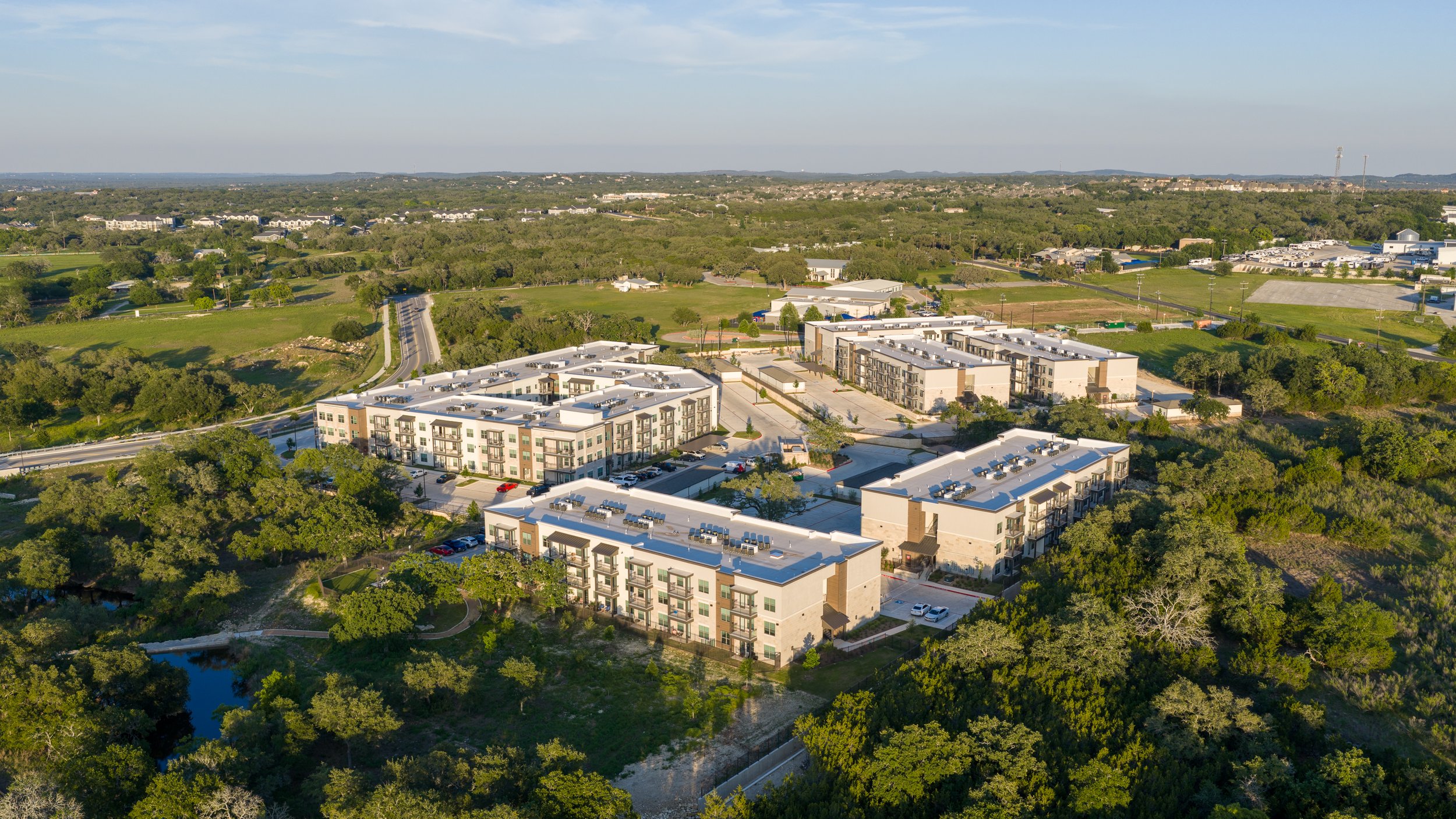 Aerial view of a residential area with modern apartment buildings surrounded by greenery, trees, and open fields under a clear sky.