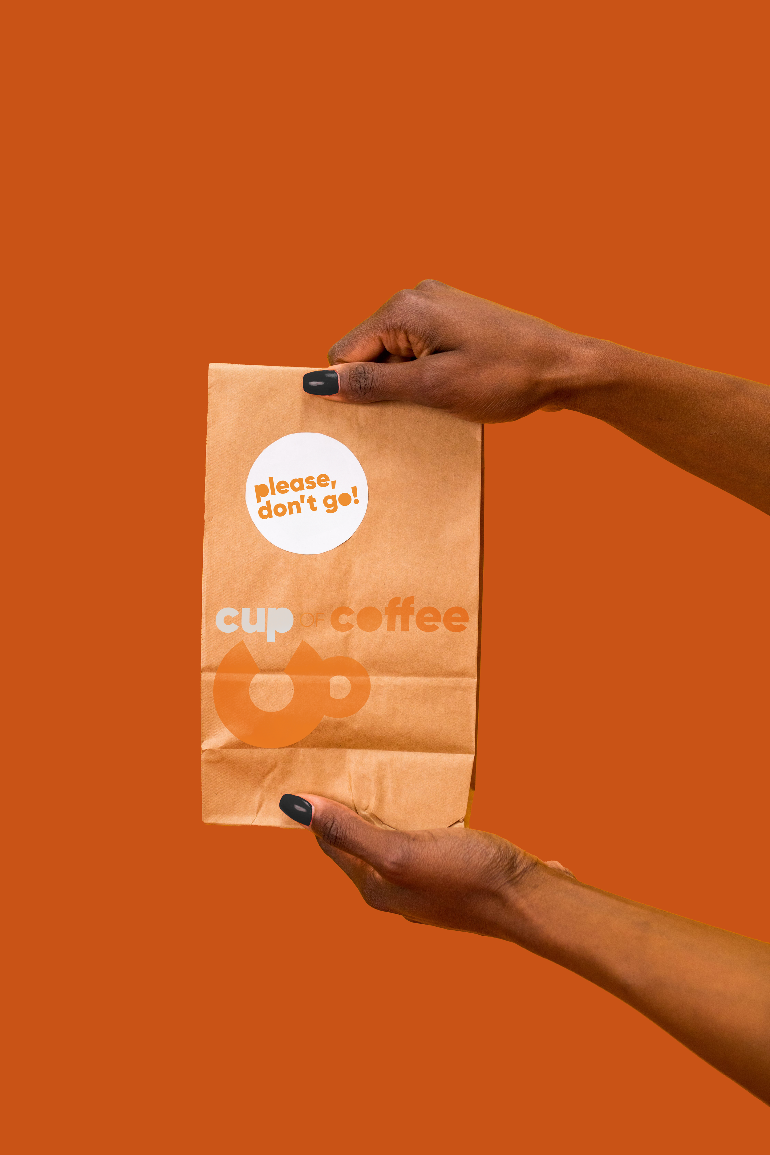Hands holding a brown paper bag with a white circle sticker that says 'please, don't go!' and the words 'cup of coffee' printed on the bag against an orange background.