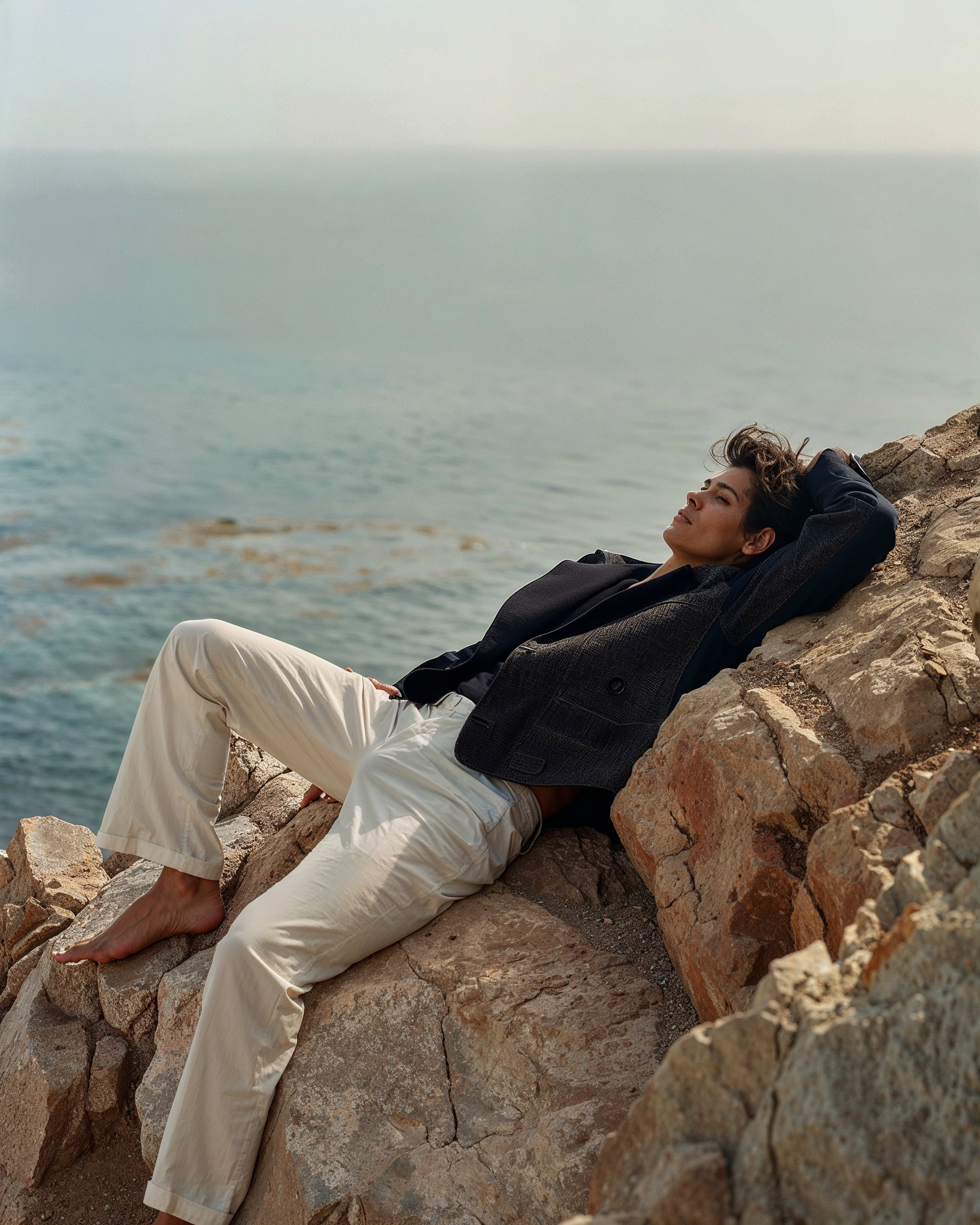 A woman relaxing on rocks near the water with outstretched legs, leaning against a large boulder, eyes closed, wearing a black blazer and cream pants.