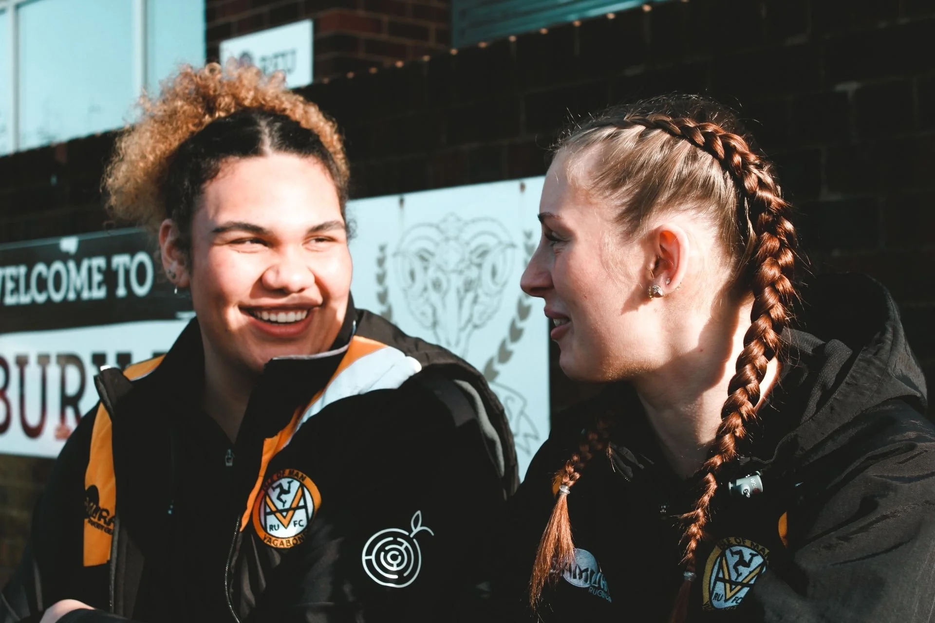 Two women with team jackets smiling and talking outdoors against a wall with signs, one with curly hair and the other with braided hair.